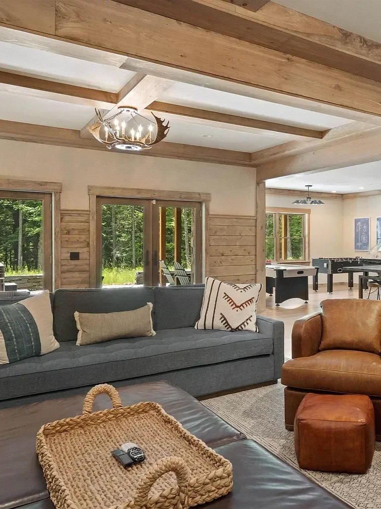 Living room with a couch, armchair, and coffee table, with wooden interior, large windows, and a view of a wooded outdoor area.