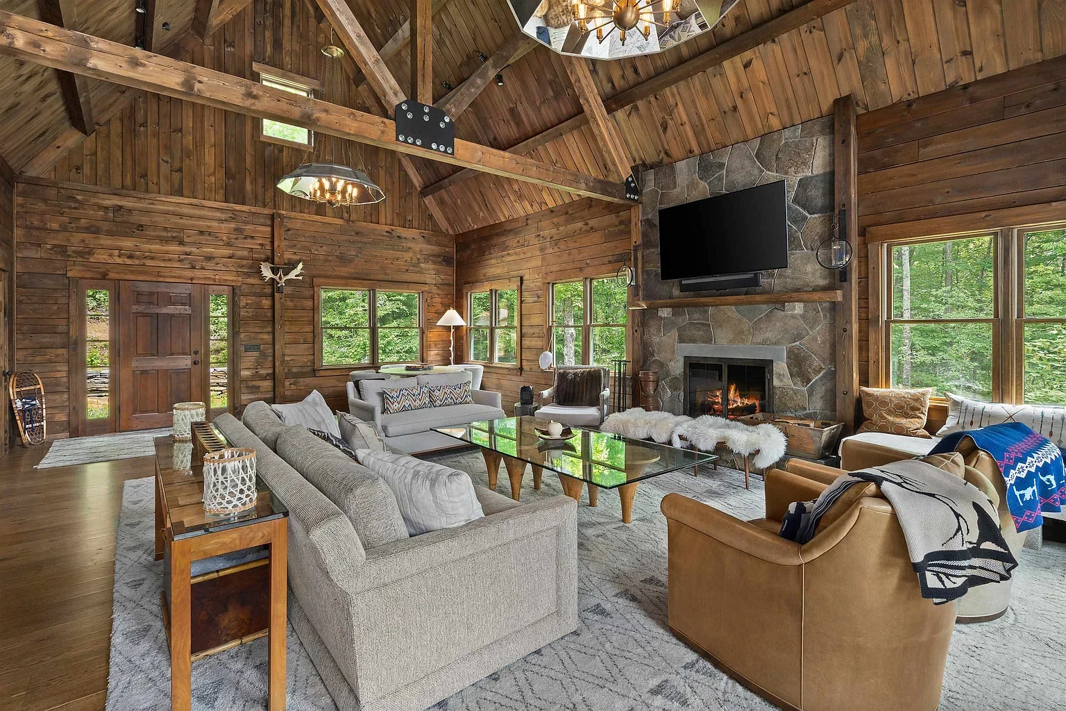 Living room with wooden walls and ceiling, a stone fireplace with a mounted TV, multiple windows showing trees outside, and a variety of sofas and chairs surrounding a glass coffee table.