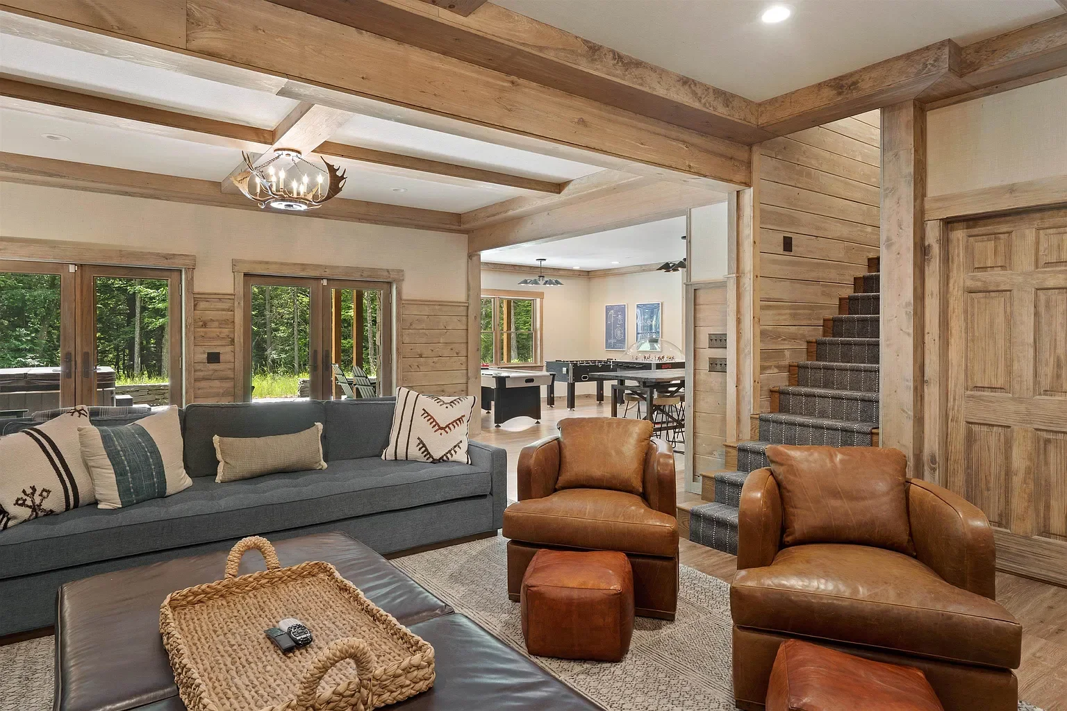 Living room with wood-paneled walls and ceiling trim, featuring a gray sofa with pillows, leather armchairs, a basket on a coffee table, sliding glass doors leading outside, and a view into an open kitchen and game room area with a ping pong table an