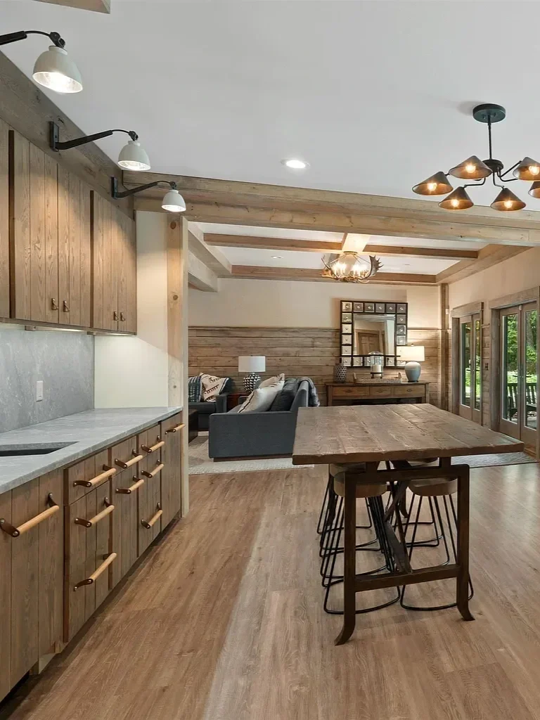 Open-concept kitchen and living room with wooden accents, including cabinets, wall paneling, and ceiling beams. There is a large wooden dining table with barstools, a gray sofa with patterned pillows, and modern lighting fixtures.