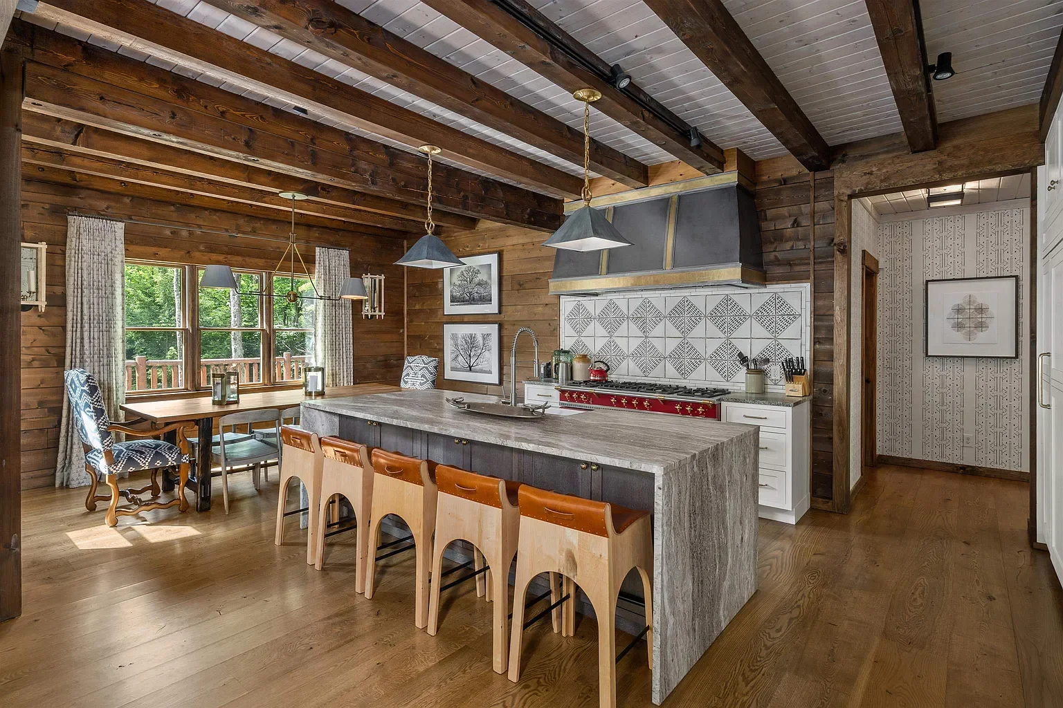 A rustic kitchen with wood-paneled walls and ceiling, an island with a gray stone top, and a red stove. A dining area with a wooden table, upholstered chairs, and a large window with curtains overlooks a green outdoor view.