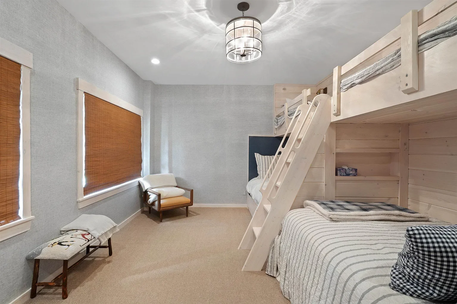 A bedroom with beige carpet, light gray textured walls, and three windows with brown blinds. There is a white bunk bed with a ladder, a striped bedding on the lower bed, and a pillow. A navy headboard is visible behind the upper bunk. To the left, th