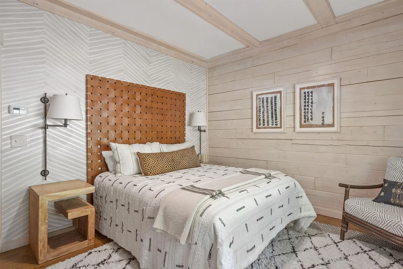 A bedroom with a bed that has a wooden panel headboard, white and striped bedding, and decorative pillows. There are wall-mounted lamps on either side of the bed, a wooden nightstand, a patterned armchair with pillows, and framed artwork on a light wooden wall.