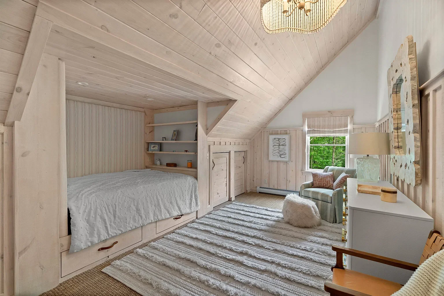 Cozy bedroom with a sloped ceiling, white wooden walls, a bed with a pull-out drawer, shelves with decor, a window showing green trees, a striped armchair with pillows, a white dresser with a lamp, and a textured rug.
