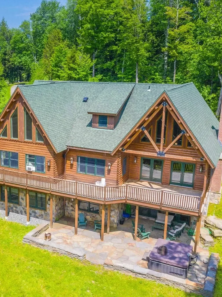 A large wooden house with a green roof, multiple windows, and a spacious deck, surrounded by green trees and a grassy lawn.