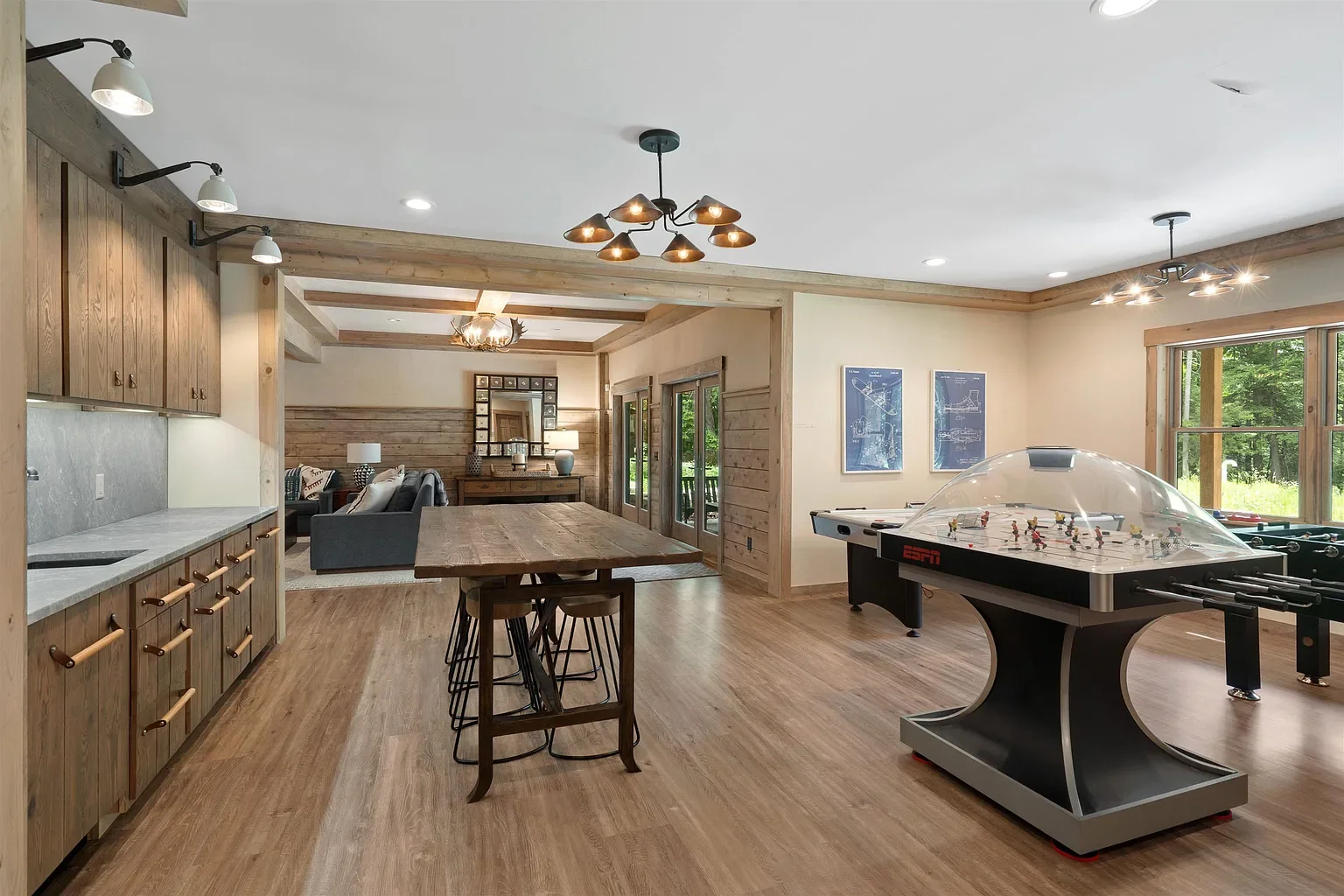 Open-concept game room with an air hockey table, foosball table, a wooden kitchen island, and a living area with a sofa and sliding glass doors leading outside. The room features wooden accents and large windows with a view of greenery.