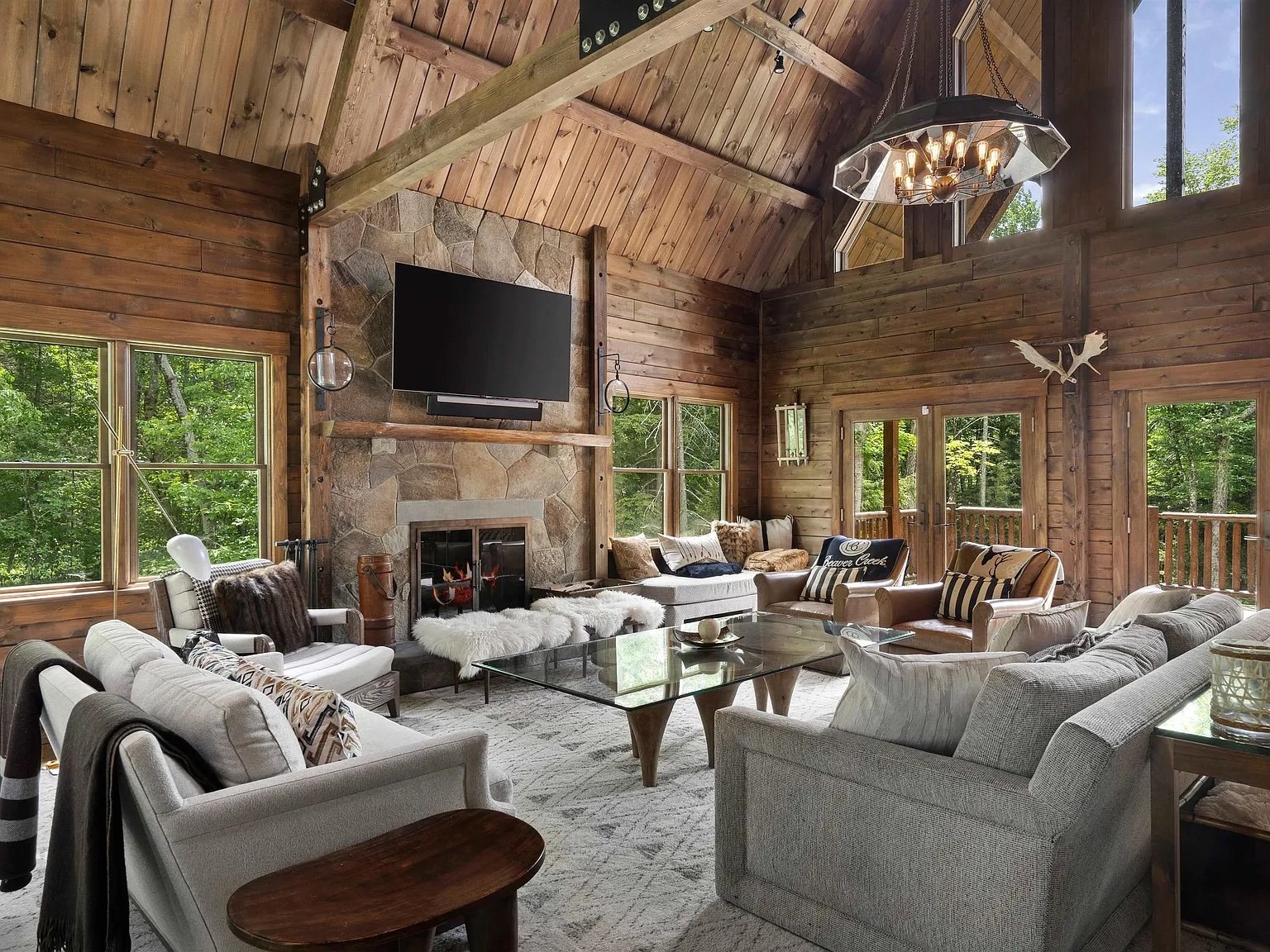 A rustic living room with wooden walls, large windows, and a vaulted wooden ceiling. It features a stone fireplace with a mounted TV above, surrounded by comfortable seating including sofas, armchairs, and a glass coffee table. Decor includes cozy th