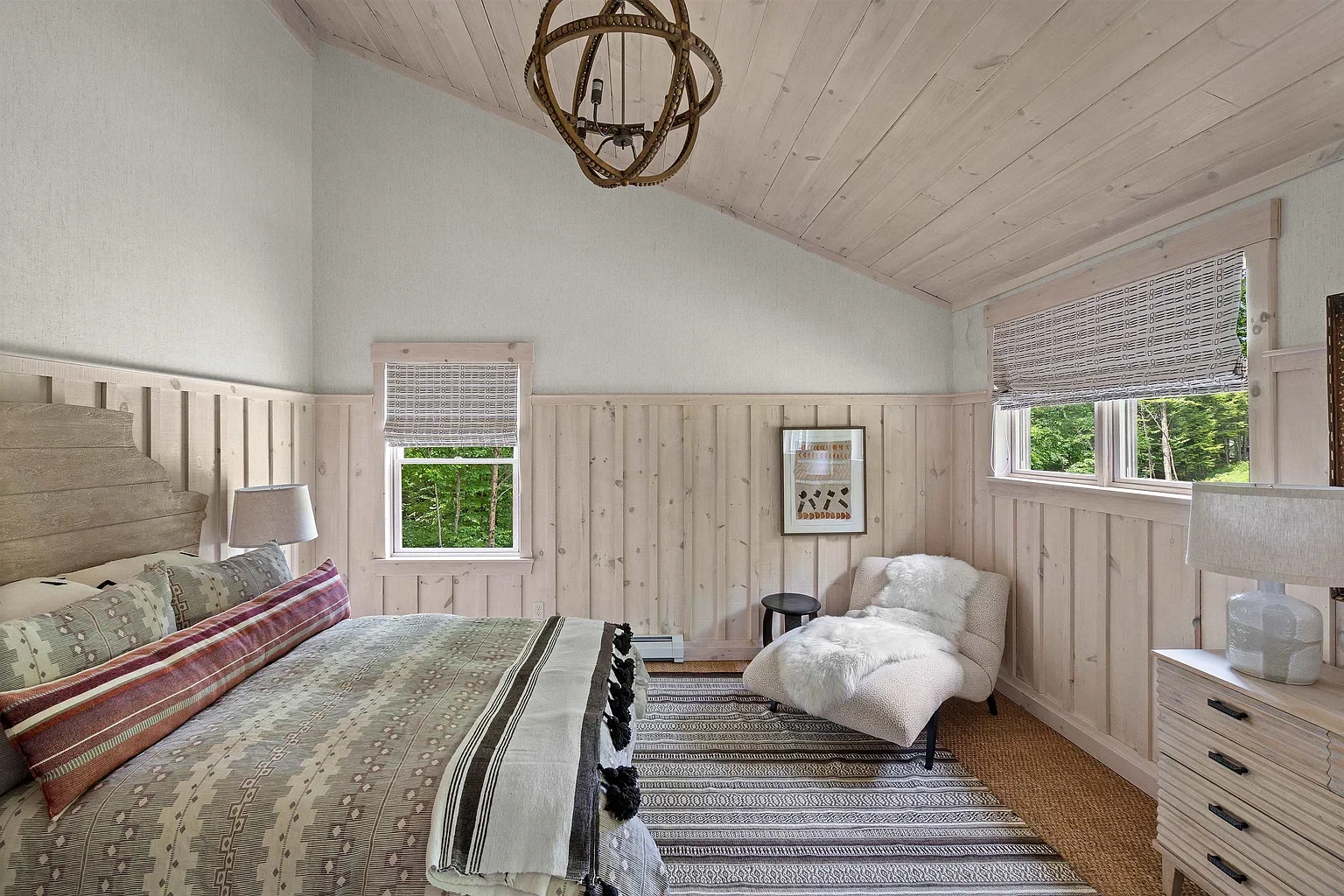A bedroom with light wood paneling on the walls and ceiling, a bed with patterned bedding, two windows with shades, a small upholstered chair with a throw, a dresser, and a striped rug.