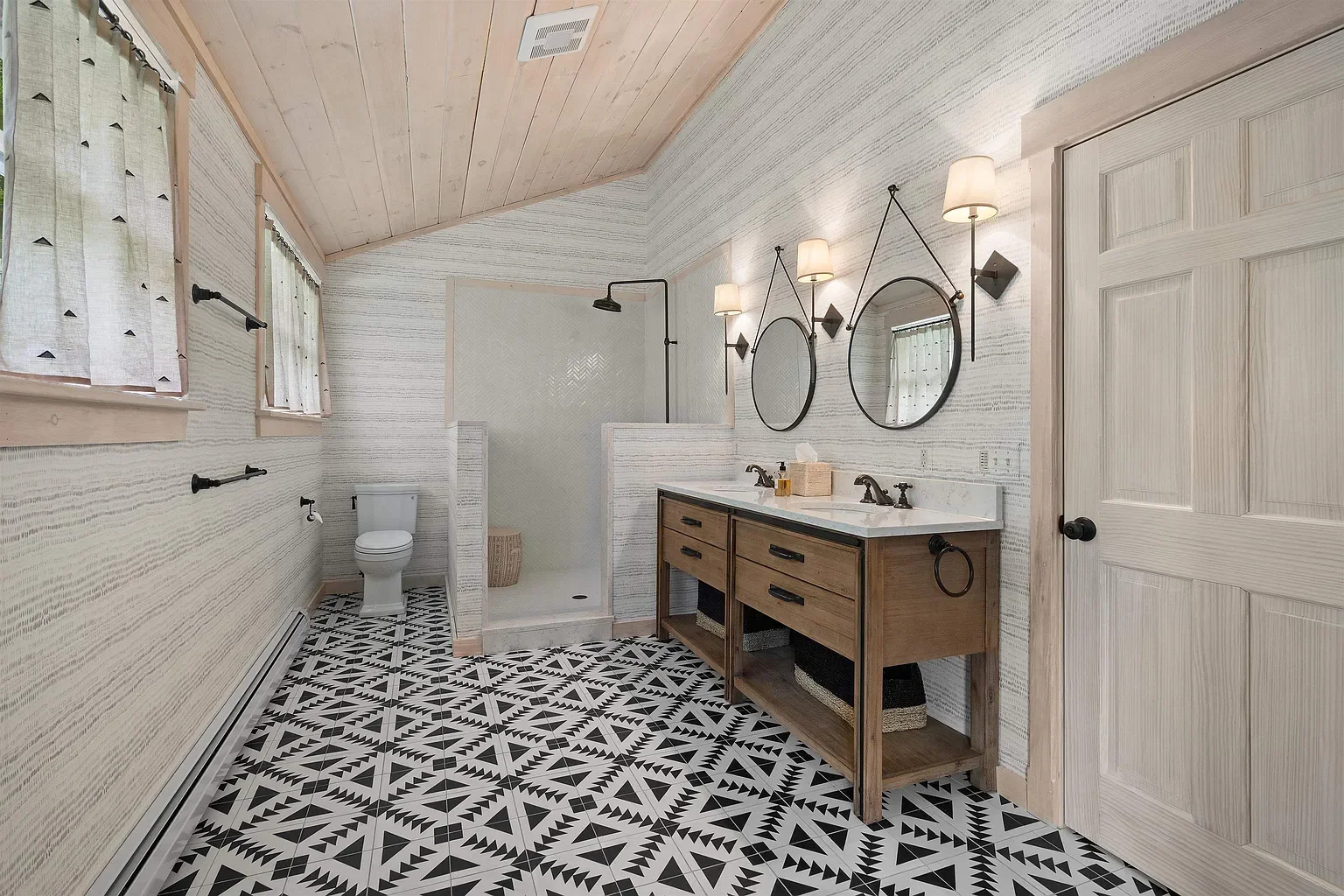 Bathroom with patterned black and white floor tiles, white and wood vanity with two sinks, two round mirrors, three wall-mounted lights, wood-paneled ceiling, shower area with black rainfall showerhead, toilet, and window with curtains.