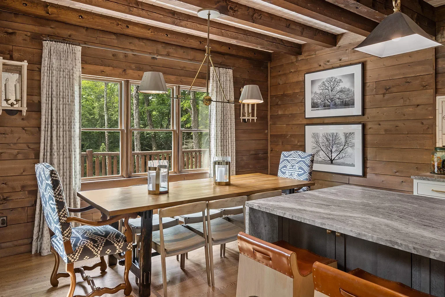 Interior of a rustic dining area with a wooden wall, large window showing trees outside, a wooden dining table with patterned chairs, and black-and-white framed photographs of trees on the wall.