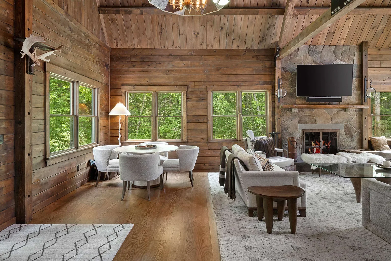 Living room with wood-paneled walls, large windows, a stone fireplace with a mounted TV, and white modern furniture, including a sitting area and a dining table, overlooking a green forest outside.