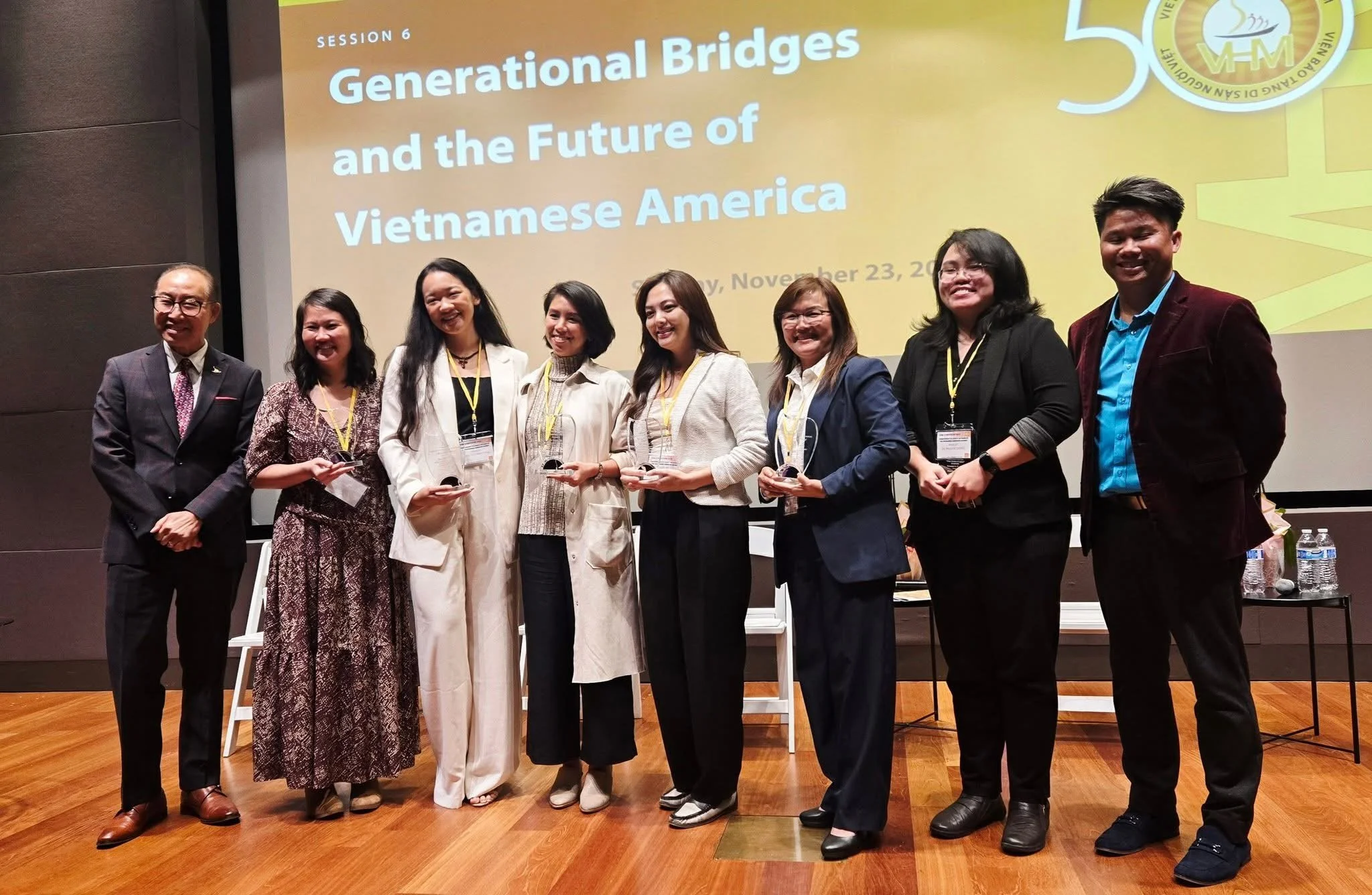 A group of nine people, including Tracy La, stand on a stage in front of a yellow presentation screen. The screen displays the title 'Session 6: Generational Bridges and the Future of Vietnamese America' and the date November 23.