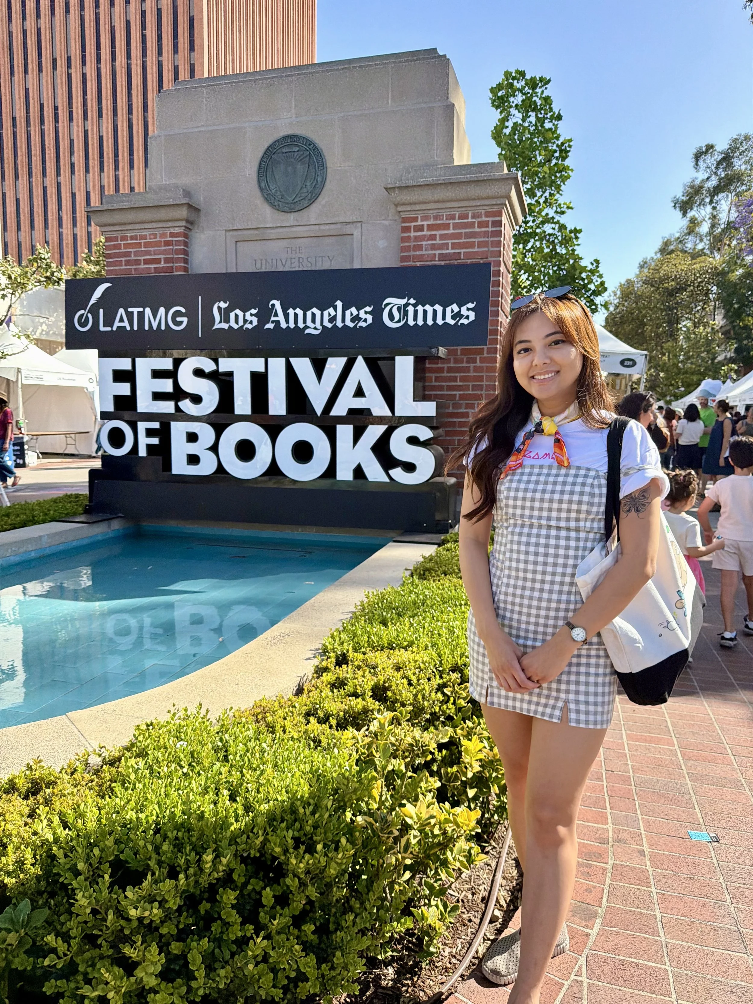 A young woman smiling outdoors at the LA Times Festival of Books at the University of California, Los Angeles. She has long brown hair, wears sunglasses on her head, a checkered dress, and sneakers. There are tents and people in the background, with a brick and stone university entrance sign and a water fountain nearby.