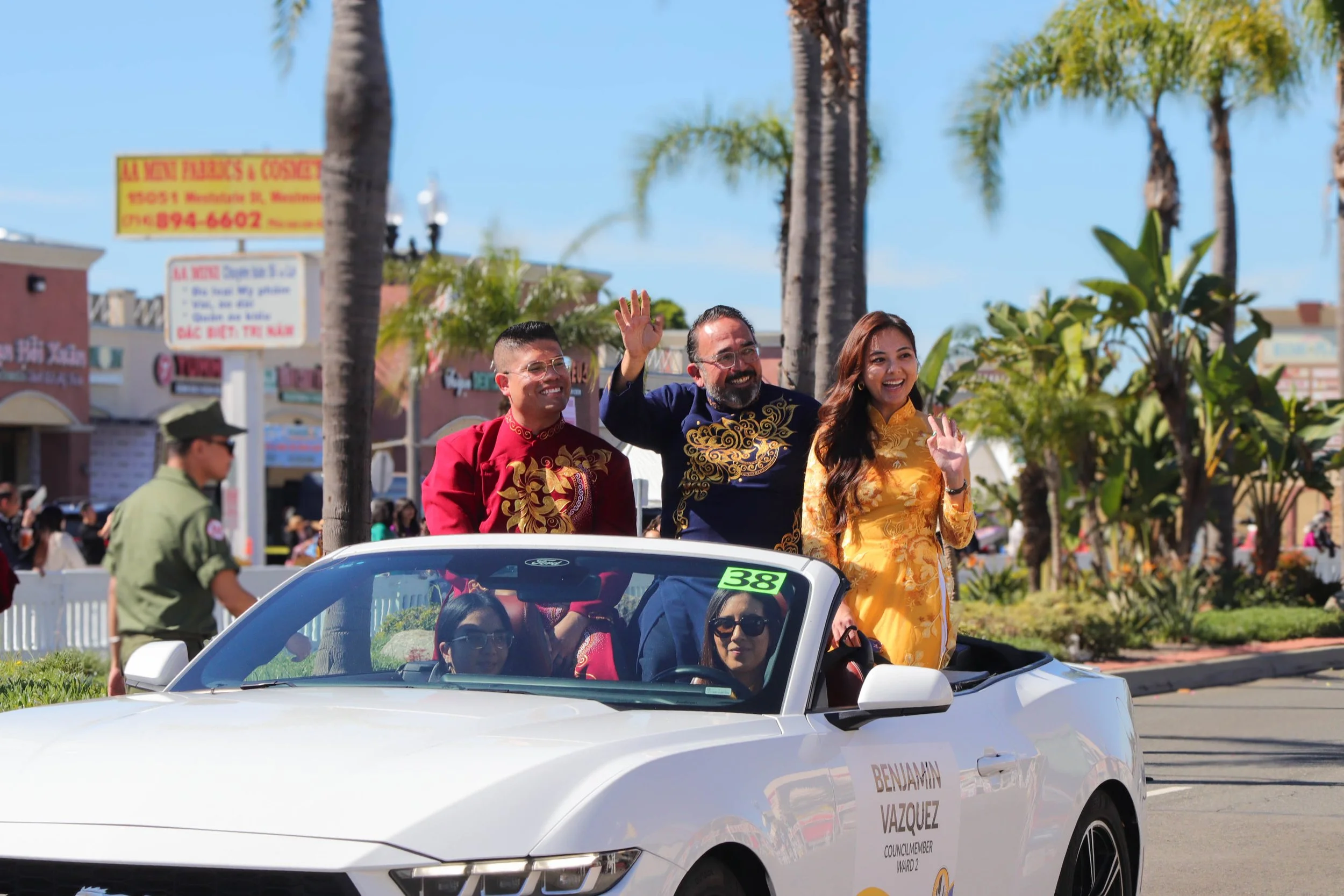 Tracy La is dressed in traditional Vietnamese clothing waving from a white convertible car during a parade. Tracy is wearing a yellow áo dài. There are palm trees and a crowd of spectators in the background.