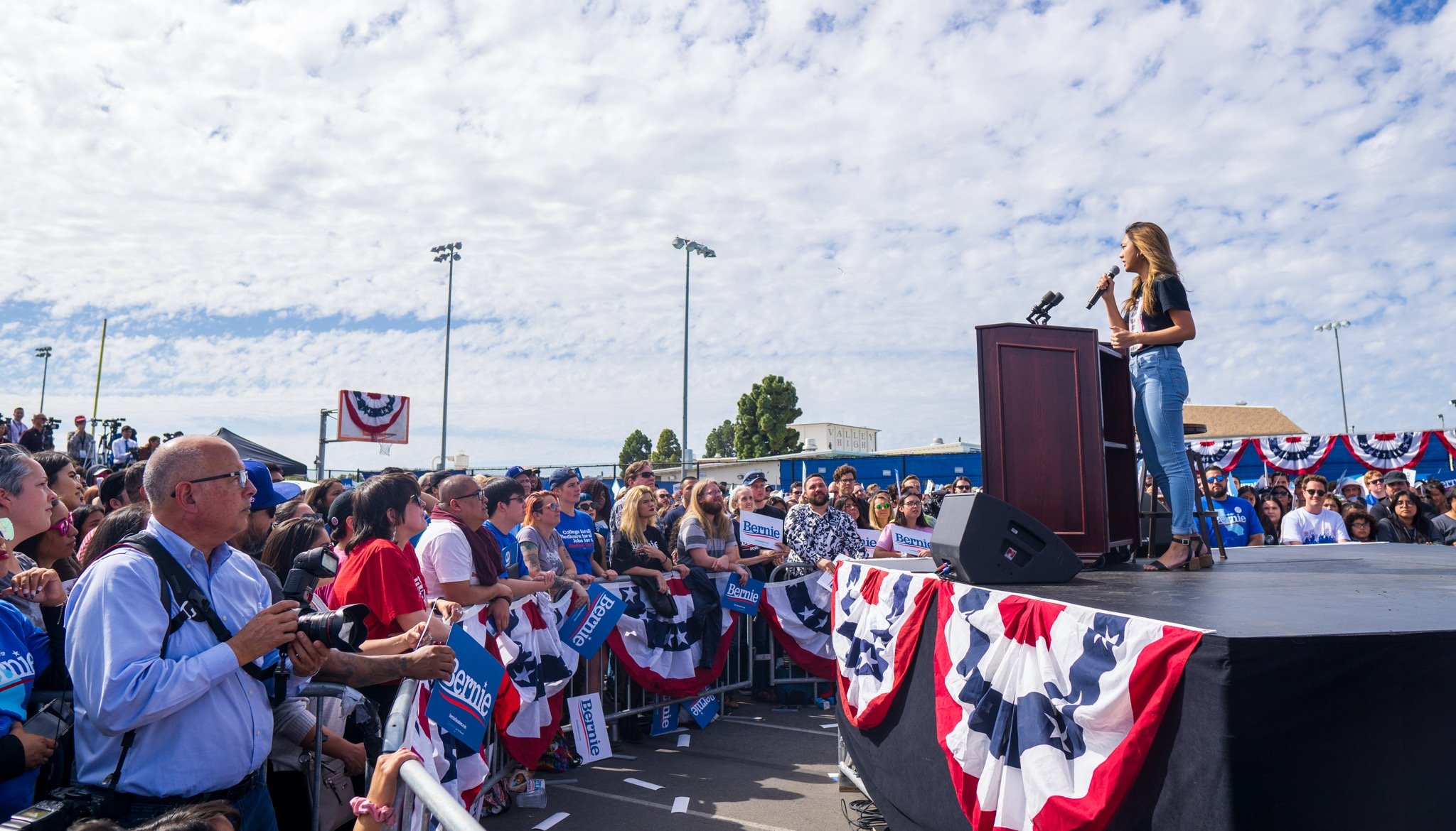 Tracy La speaks at a political rally stage with a microphone, crowd seated and standing in front, holding signs that say 'Bernie,' outdoors on a partly cloudy day, decorated with red, white, and blue banners.