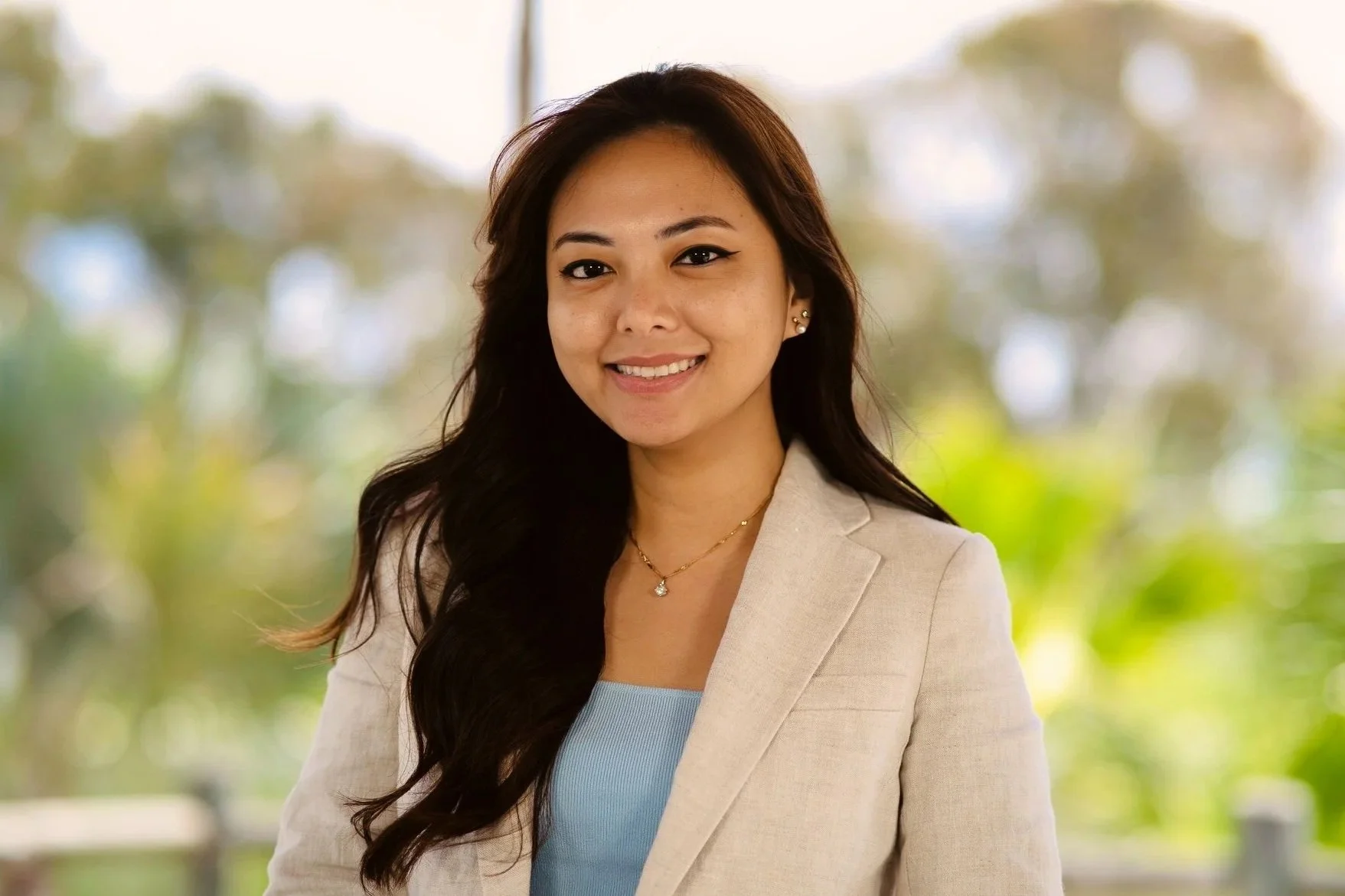 Portrait of a smiling woman with long dark hair, wearing a beige blazer, a light blue top, and jewelry, standing outdoors with trees in the background.