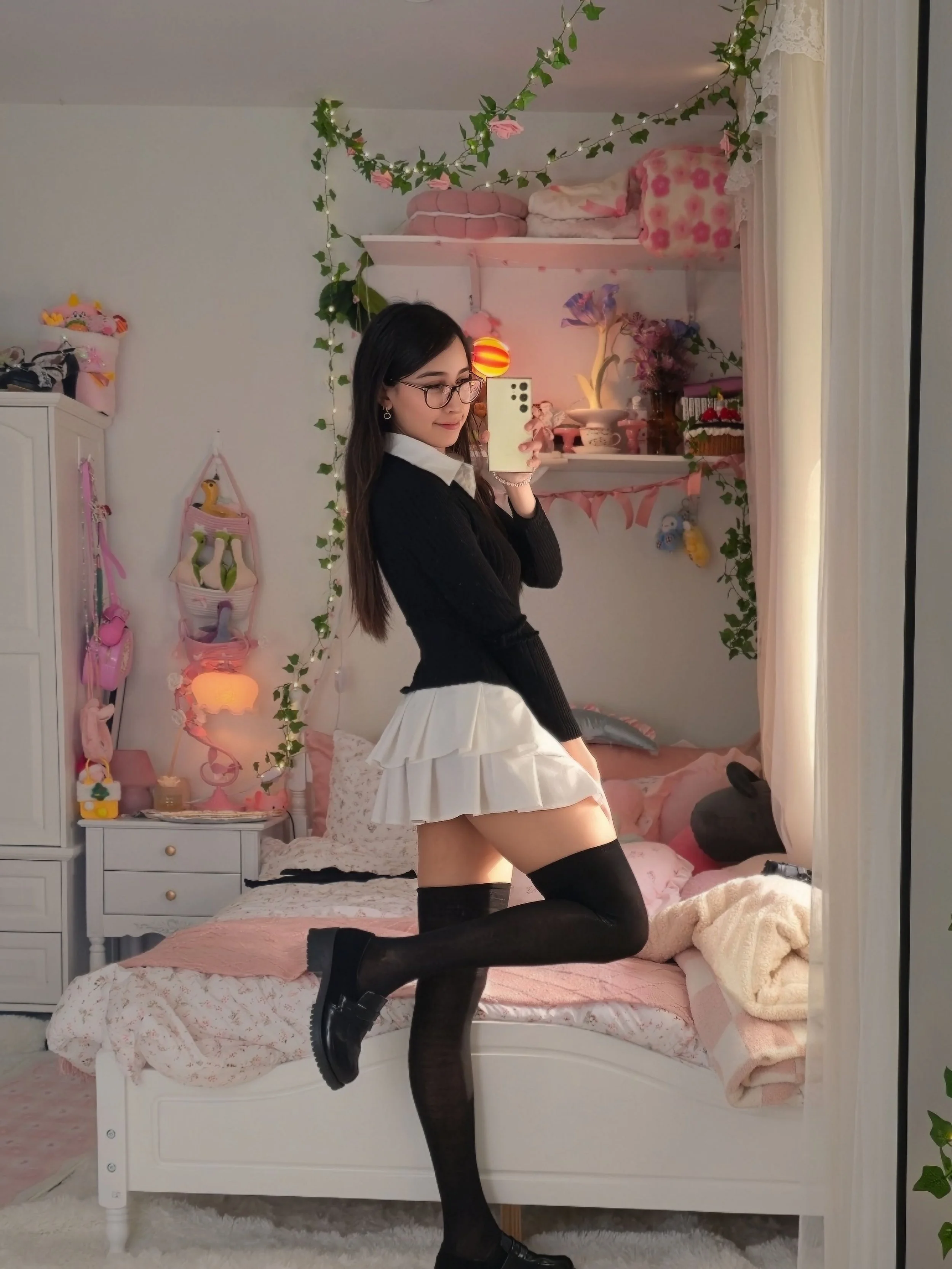 A woman with long dark hair and glasses taking a mirror selfie in a bedroom with pink and white decor. She is wearing a black sweater, white skirt, and black thigh-high stockings. The bedroom has a bed with pink bedding, a nightstand with a pink lamp, and shelves with various decorations and plush toys. Green ivy garland and fairy lights are hanging across the wall.
