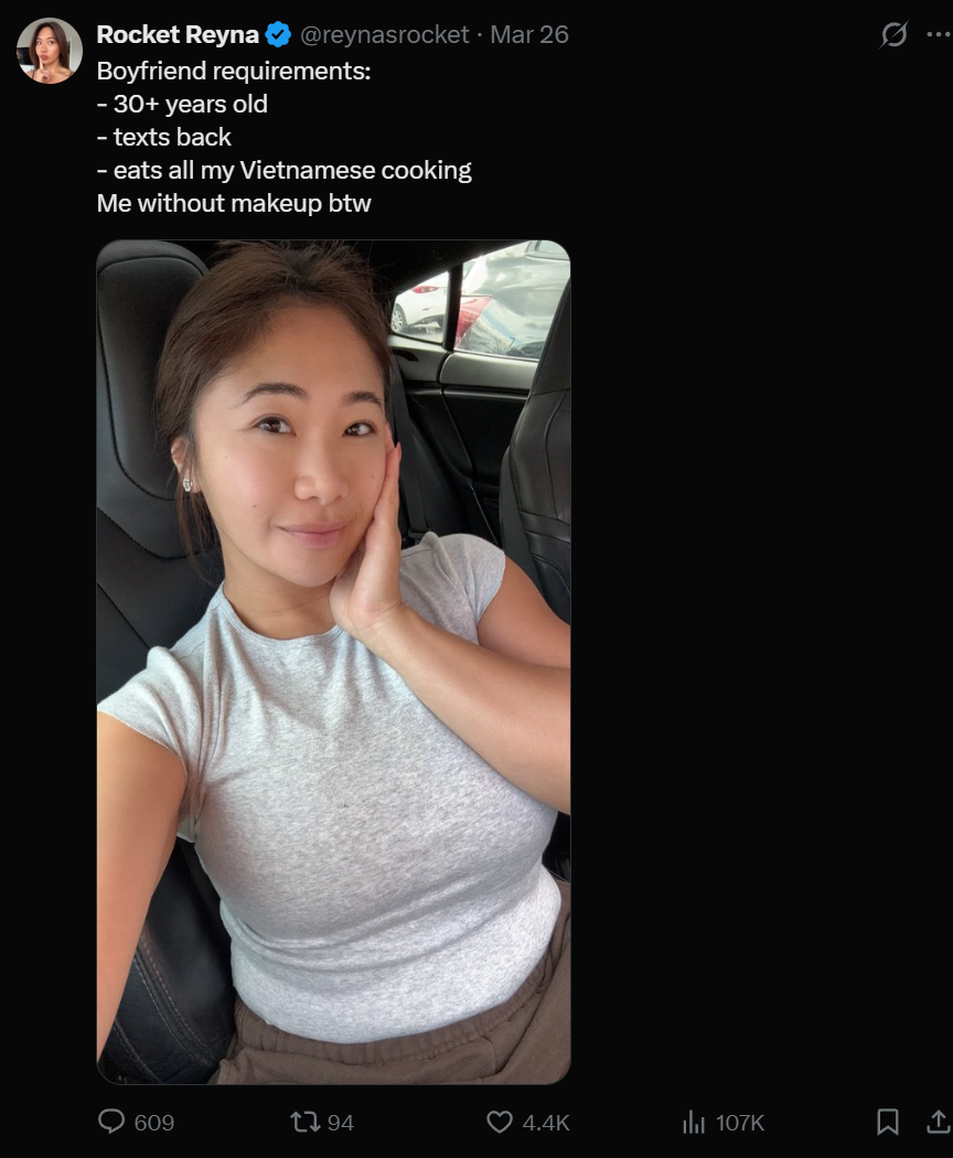 A woman sitting in a car, wearing a light gray t-shirt and brown pants, with her hand resting on her face, smiling softly for a selfie.