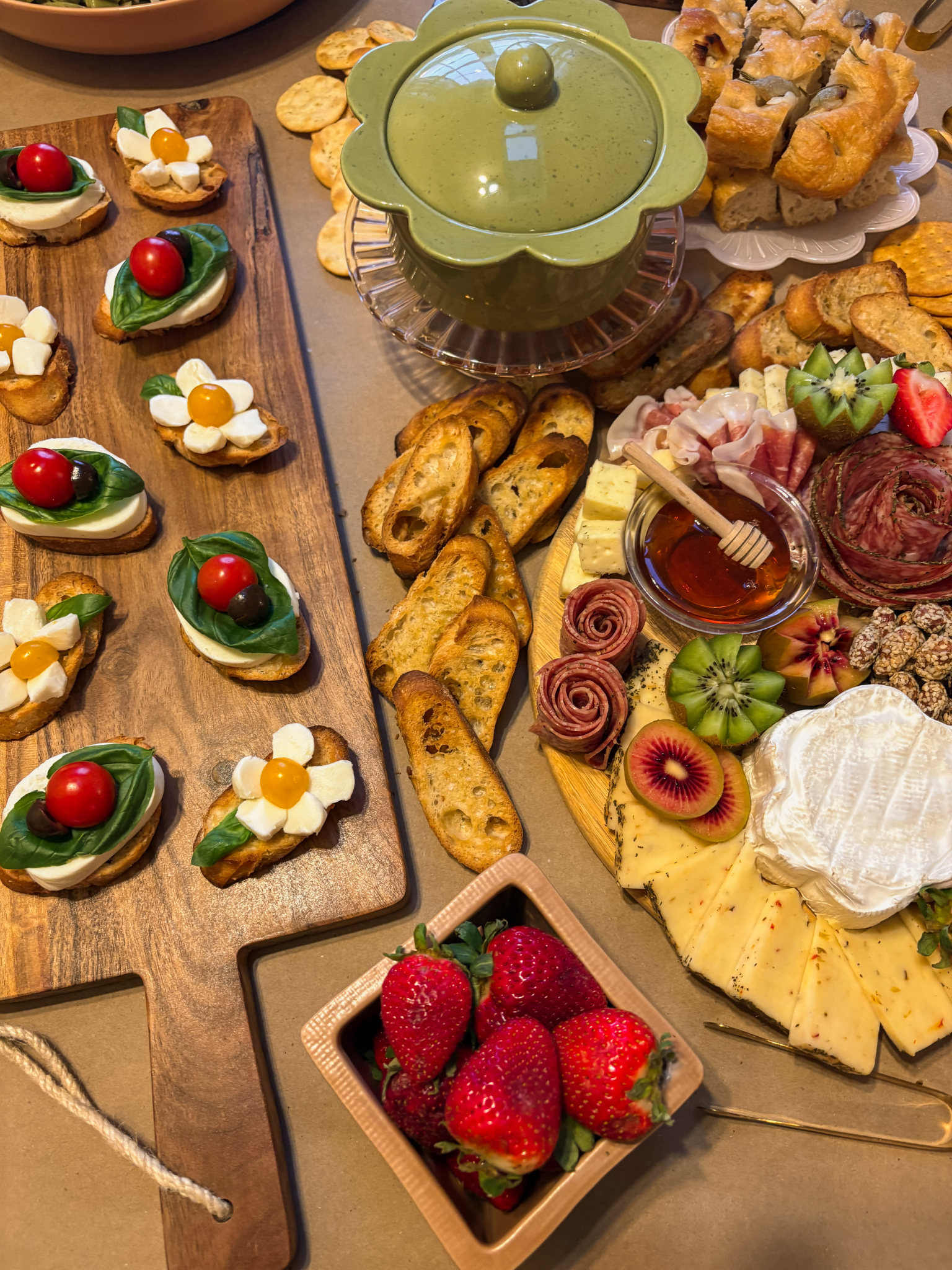 Assorted appetizers including cherry tomato and mozzarella bites, slices of baguette with toppings, cheese, cured meats, fresh fruits like strawberries, kiwi, and pineapple, a bowl of honey, and a jar of green sauce, on a table.