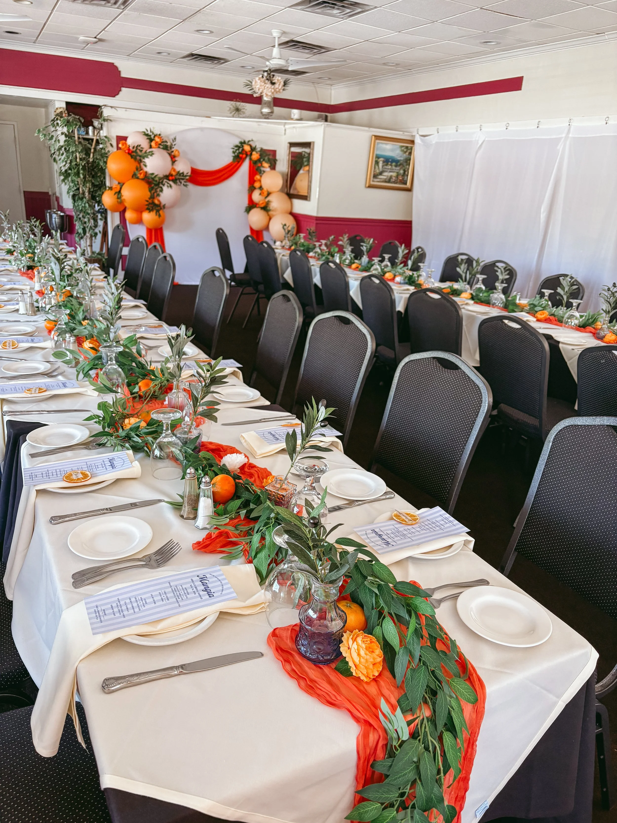 A banquet table set for a celebration with white tablecloths, decorated with orange and green foliage, orange flowers, and orange balloons on a white backdrop with drapes and balloon arrangements.
