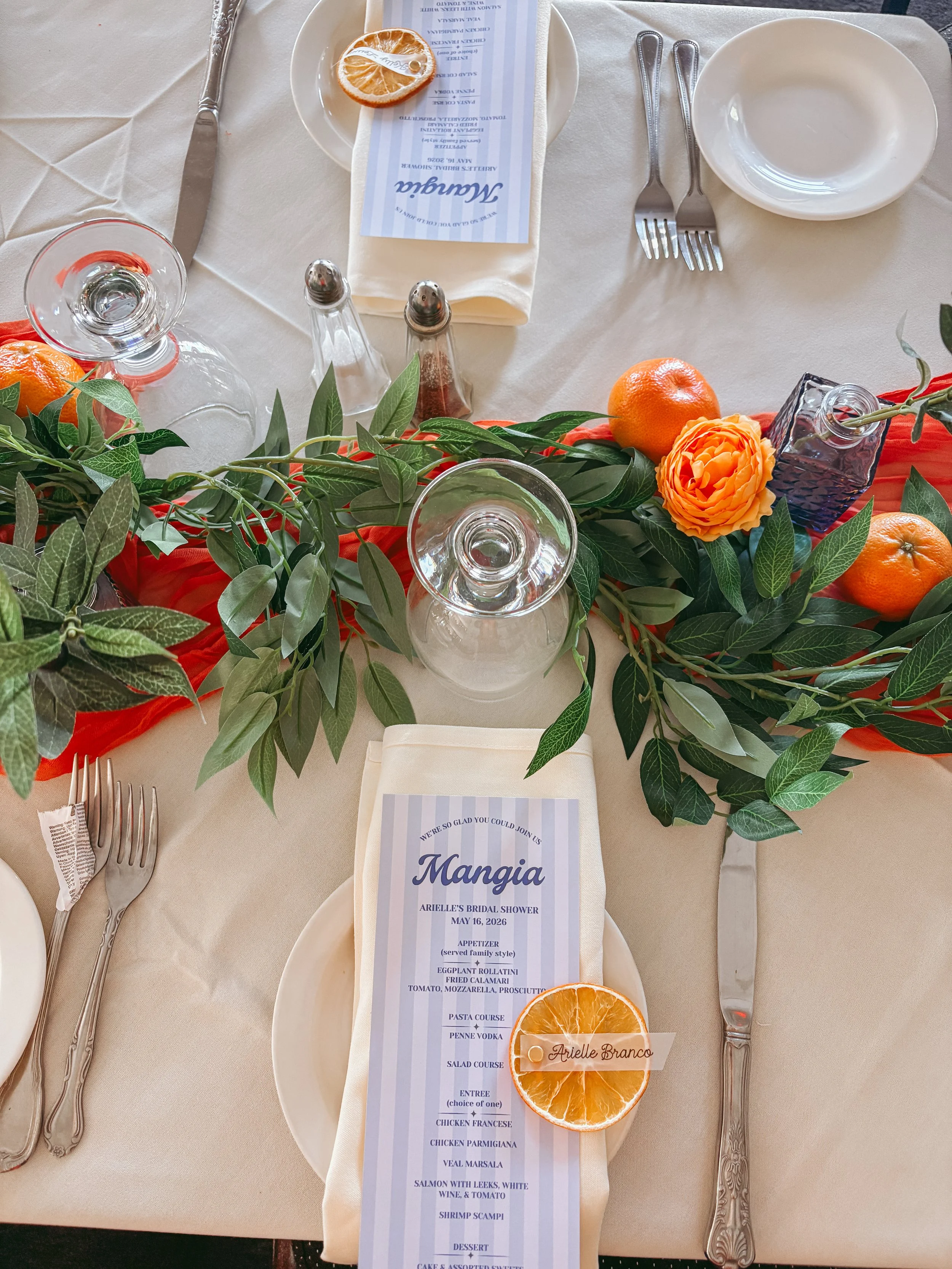 A formal banquet table setting with a white tablecloth, a menu napkin with a slice of orange, a menu card, two forks, a knife, a water glass, a wine glass, salt and pepper shakers, and decorative greenery and orange flowers. There are also whole oran