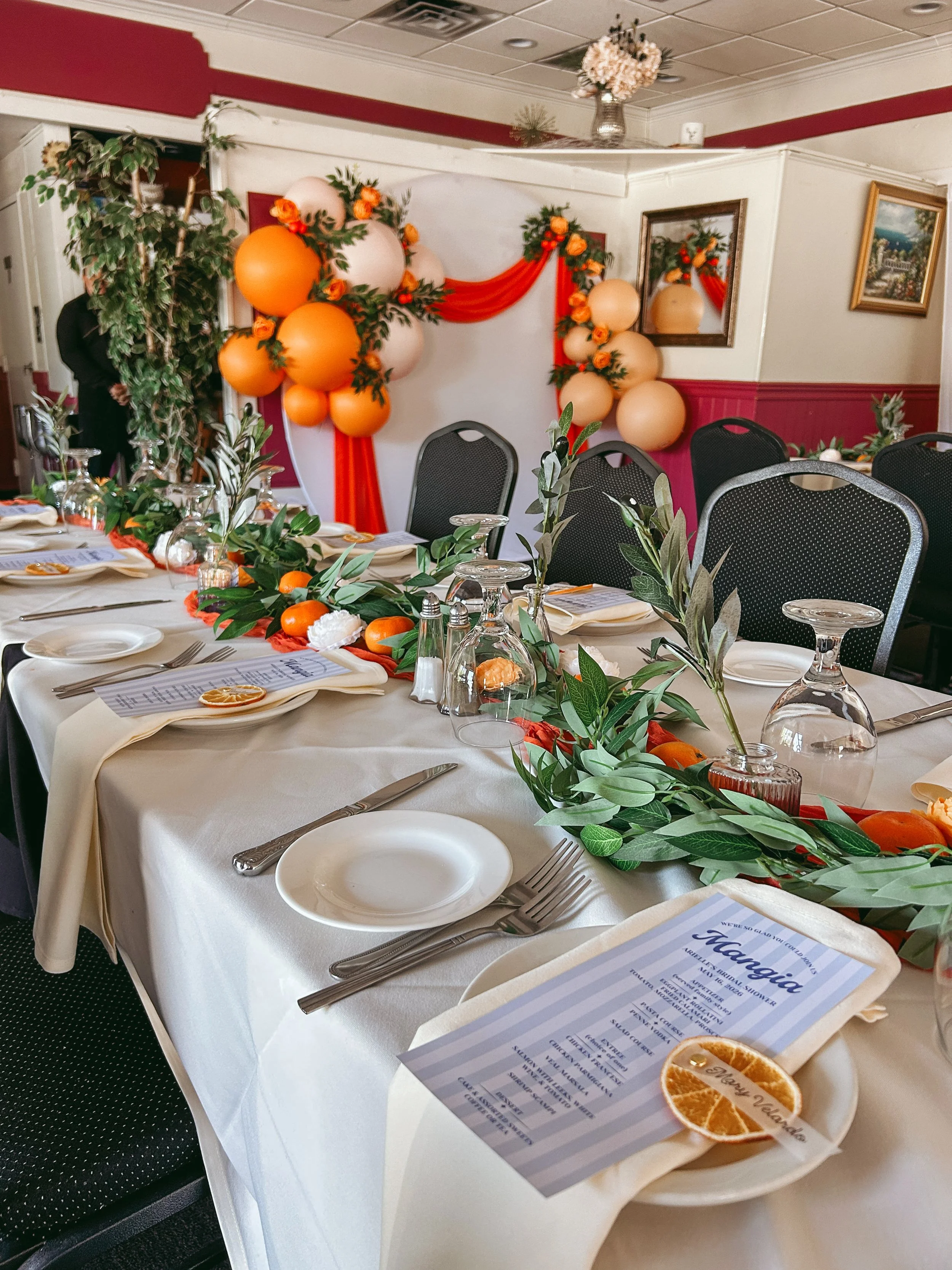 A decorated event dining table with a floral and balloon arrangement in the background, set with white tablecloth, plates, glasses, napkins, and utensils, with a menu card and orange slice on each plate, and leafy centerpieces.