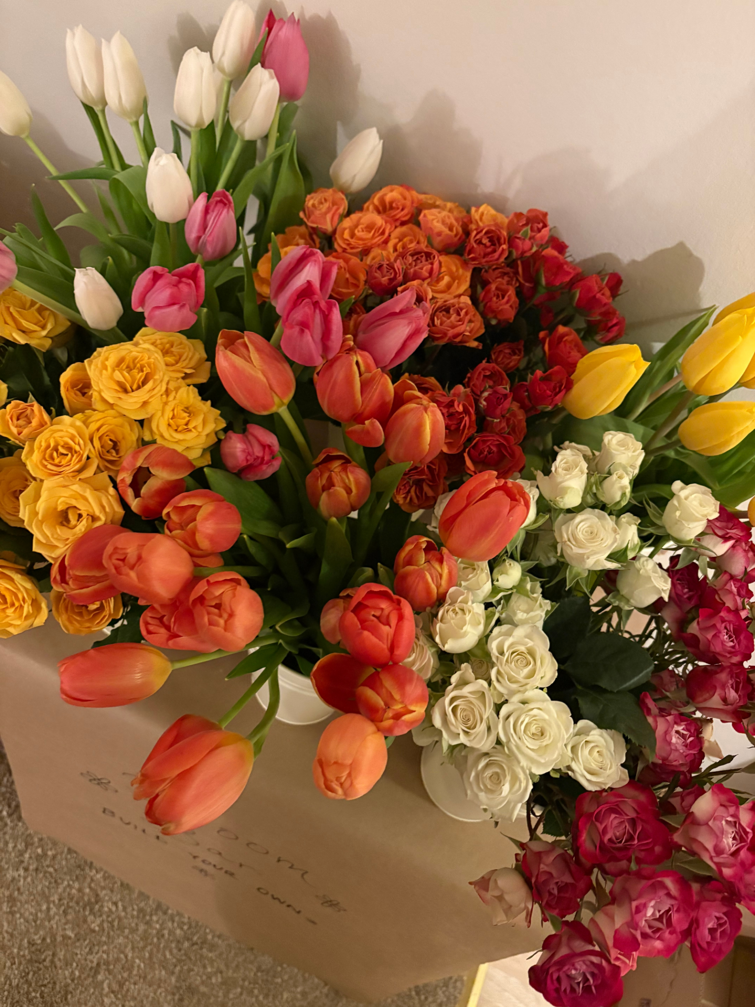 A bouquet of various colorful flowers including white, pink, orange, yellow, and red tulips and roses, arranged in a white paper wrapping.