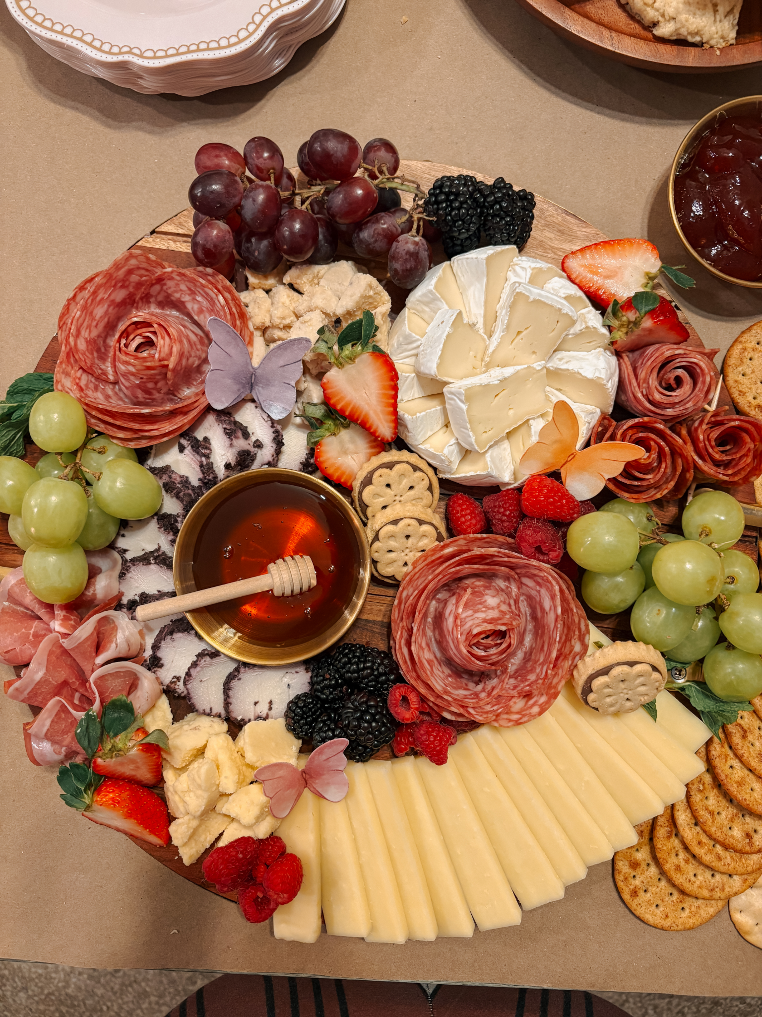 A cheese and charcuterie platter with grapes, strawberries, blackberries, raspberries, slices of cured meats, various cheeses, crackers, and honey in a small bowl, decorated with butterfly-shaped food picks.