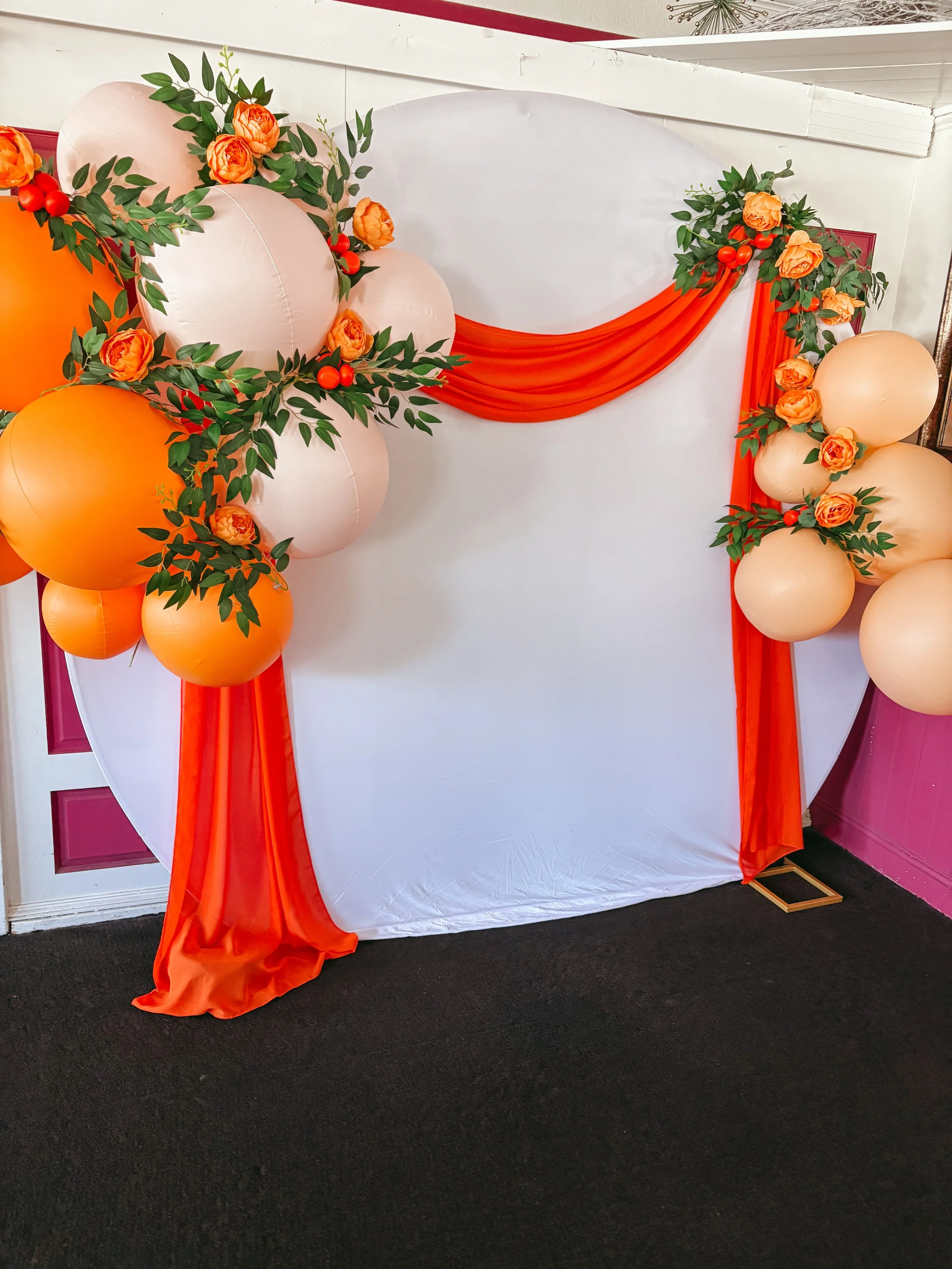 Decorative backdrop with orange and cream balloons, orange flowers with green leaves, and draped orange fabric, set against a white wall.