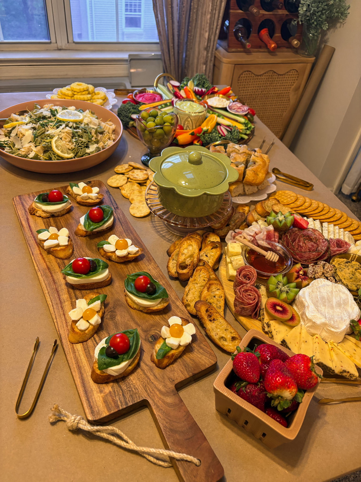 A table with a variety of appetizers, salads, sliced bread, cheese, cured meats, strawberries, and fresh vegetables, arranged for a gathering or party.