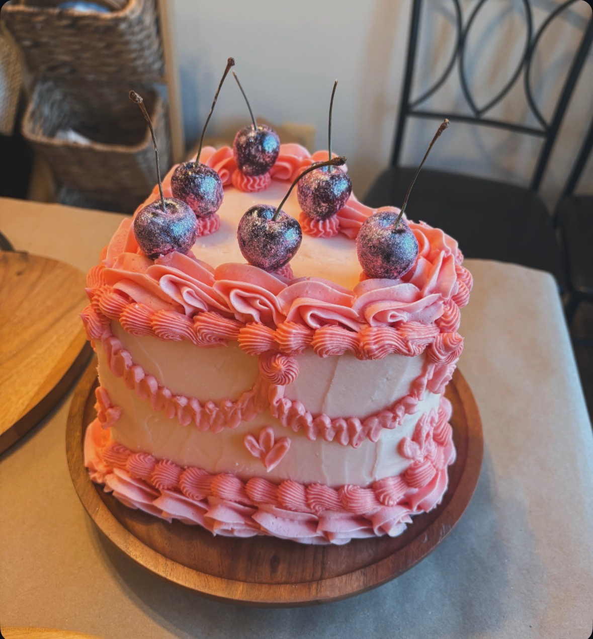 A pink and white layered cake decorated with cherries on top and pink frosting borders.