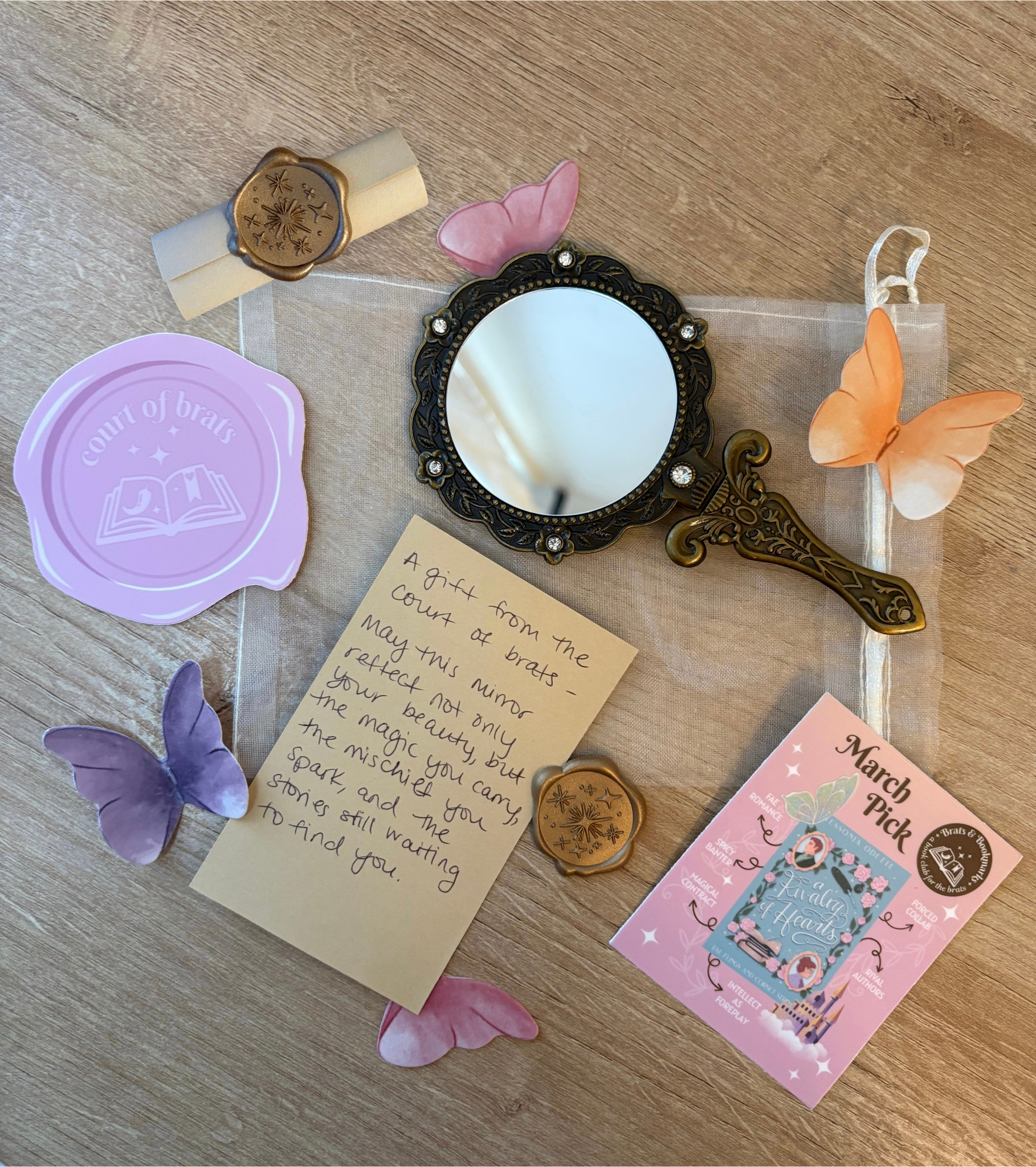 Decorative table setting with a vintage handheld mirror, pink and orange paper butterflies, a handwritten note, and promotional pamphlets for a March pick event, on a wooden surface.