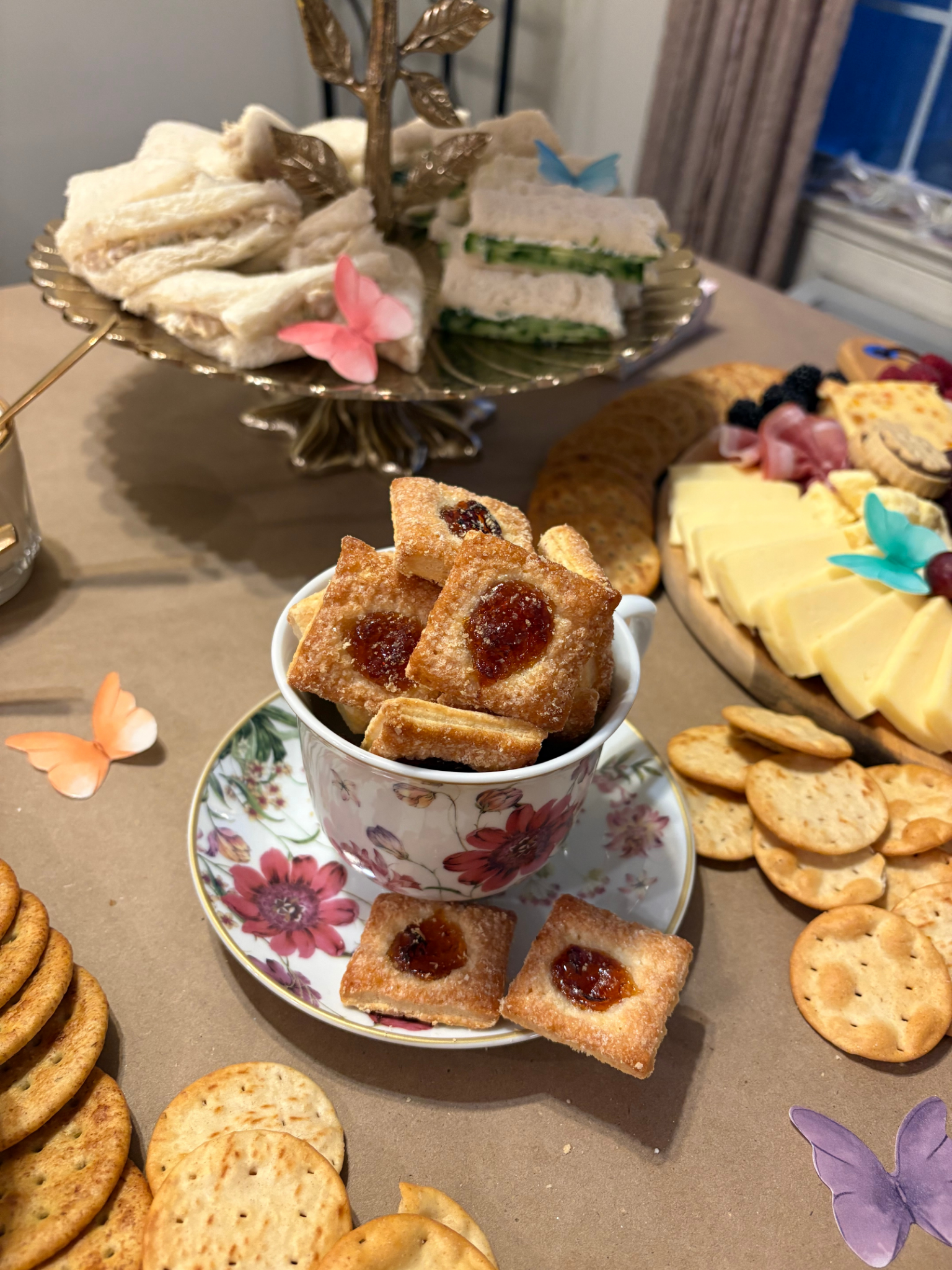 A variety of snacks and finger foods on a table, including tea sandwiches, cookies, crackers, cheeses, and decorative butterflies.