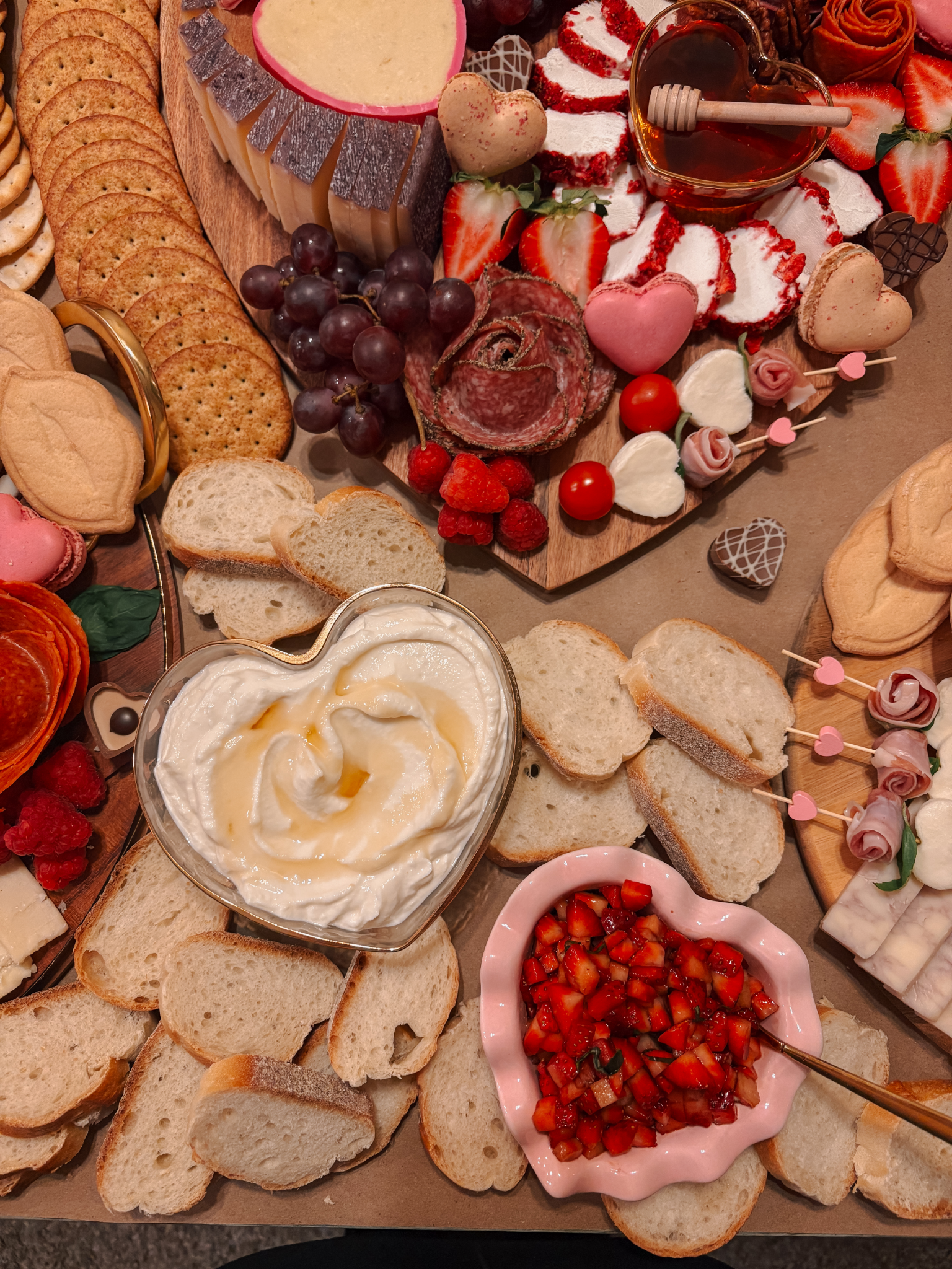 A variety of bread slices, cheeses, strawberries, grapes, raspberries, and a bowl of cream cheese with honey on top. There are also heart-shaped cookies, heart-shaped candies, and a small heart-shaped honey jar, along with some sliced meats and heart