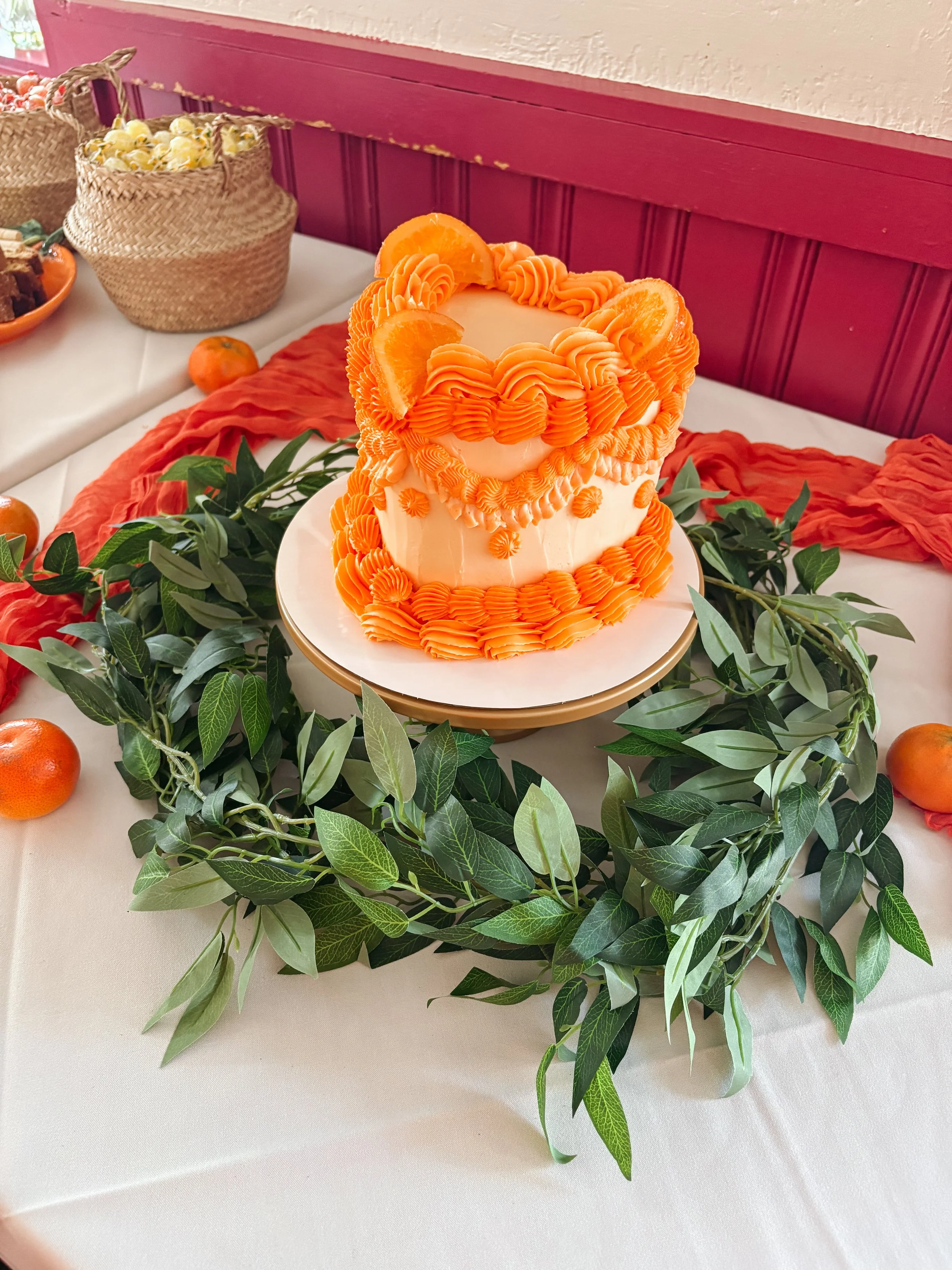 A layered cake decorated with orange frosting piped in a decorative pattern, topped with orange slices. The cake is on a white cake stand, surrounded by green leaves, on a white tablecloth.