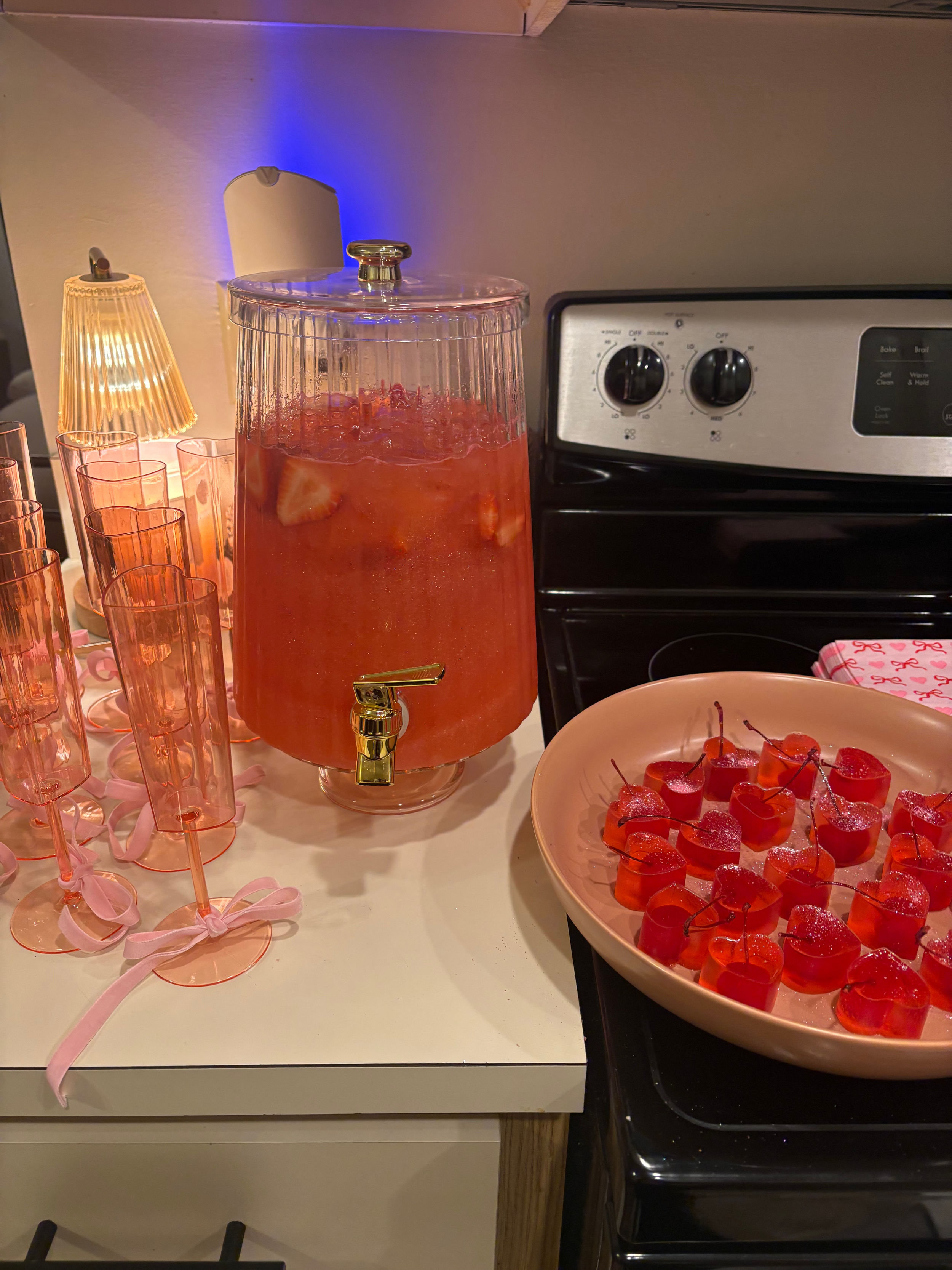 A table with a large glass dispenser with a pink drink garnished with fruit slices, pink drinking glasses with pink ribbons, a plate of cherry-shaped gummies, a small lamp, and a stove in the background.