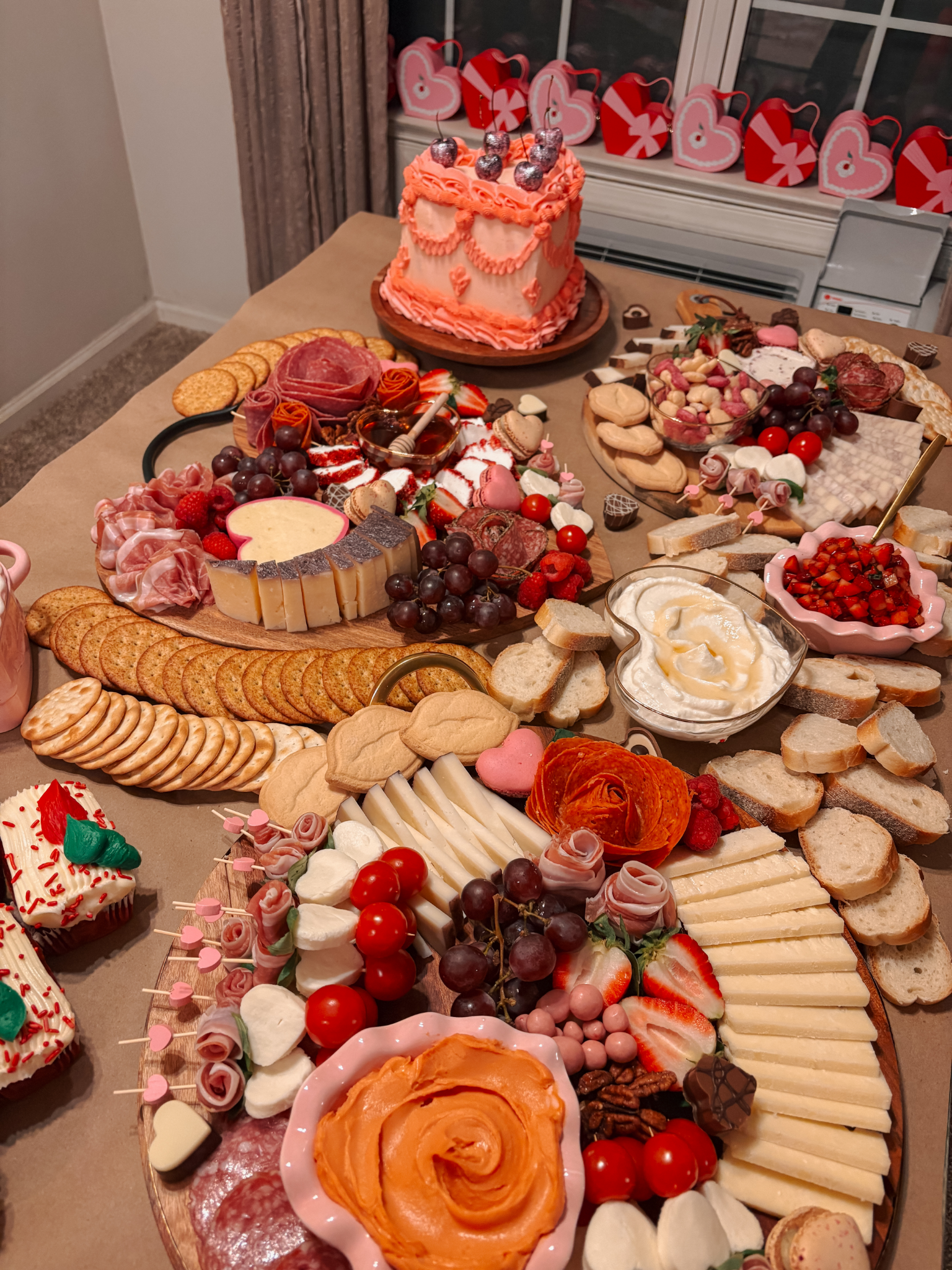 A dessert table with various sweet treats including a pink frosted cake with cherries on top, cheese, crackers, grapes, strawberries, cookies, chocolates, and heart-shaped candies, decorated for a celebration.
