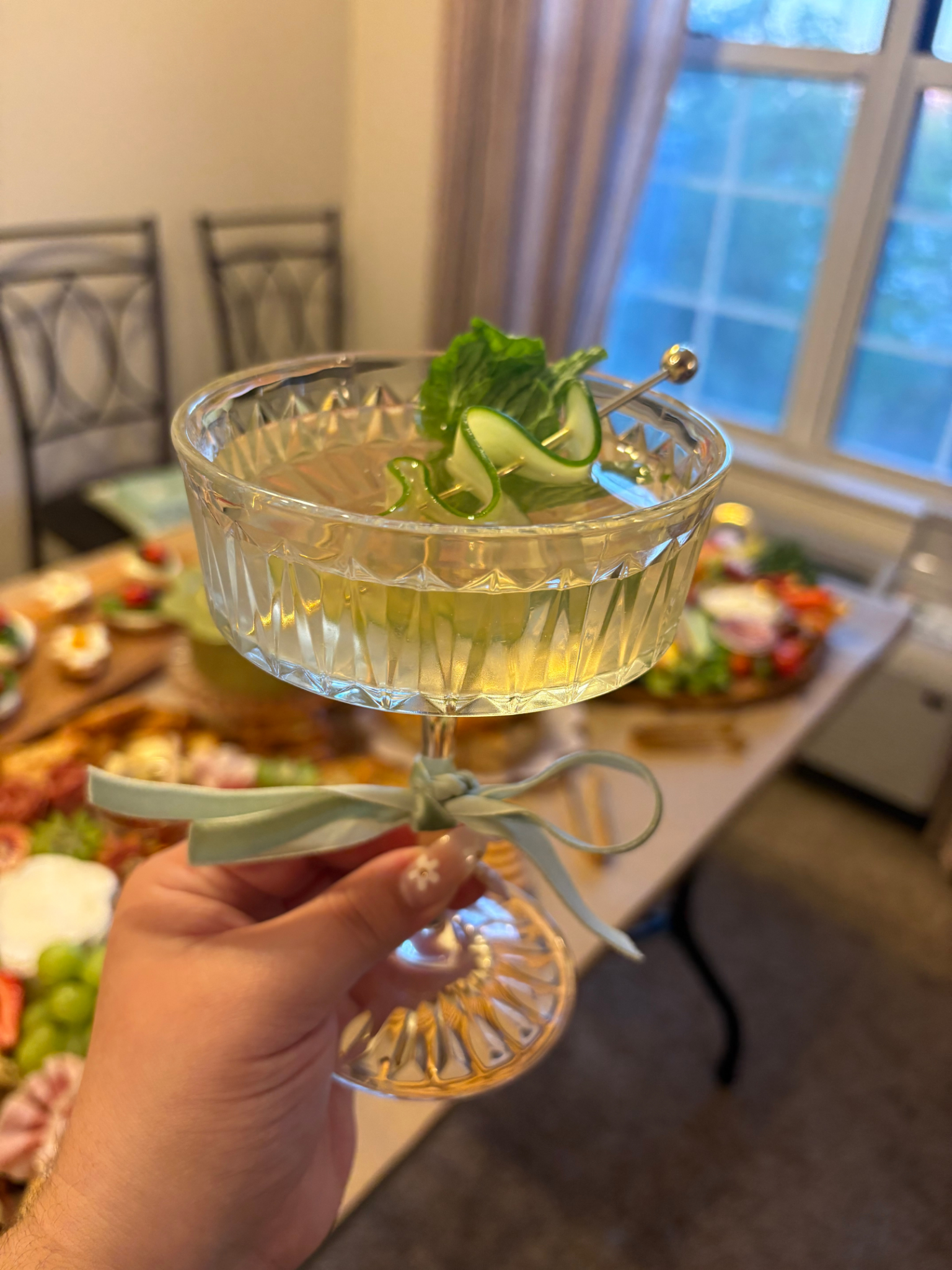 Hand holding a crystal coupe glass with a garnish of green lettuce and cucumber curls, tied with a light blue ribbon, in a dining room with a table of food in the background and a large window with curtains.