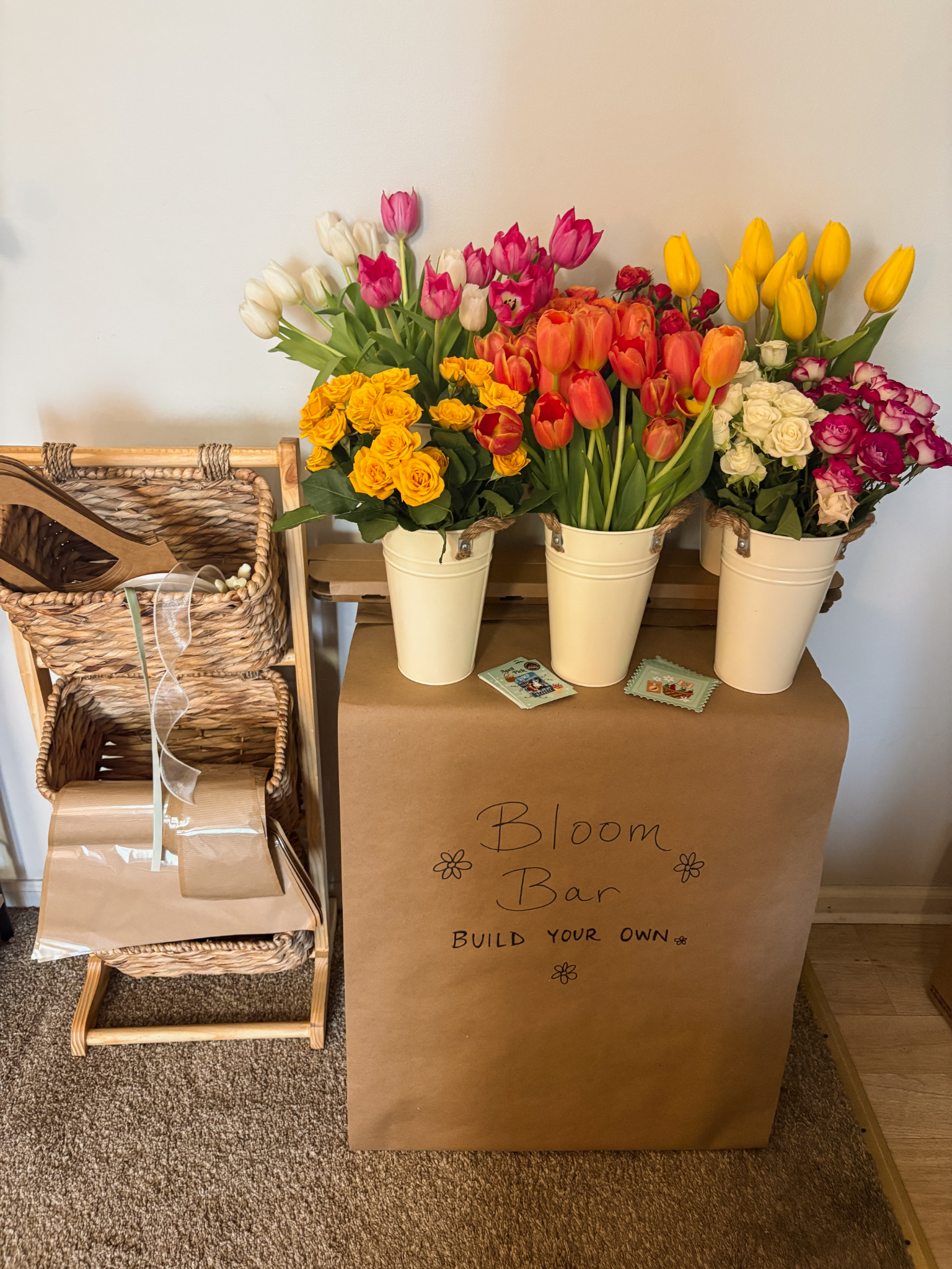 Three buckets of colorful tulips and roses on a table with a brown cardboard box labeled 'Bloom Bar Build Your Own' and some crafting supplies on a wicker shelf next to it.