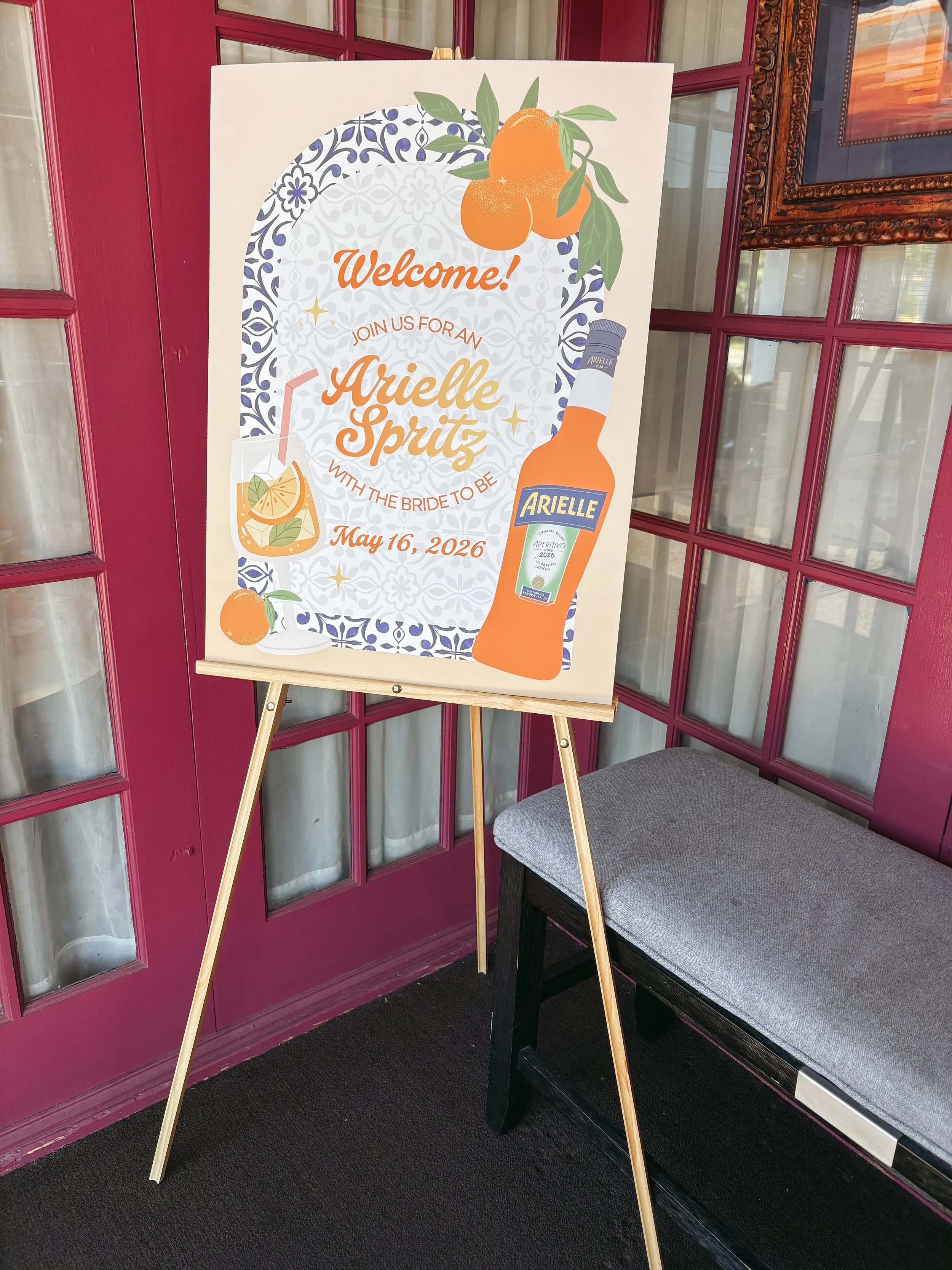 A welcome sign for a wedding event with orange citrus illustrations, a bottle of Ariel Spritz, and a glass of cocktail, placed indoors near a maroon door with windows.