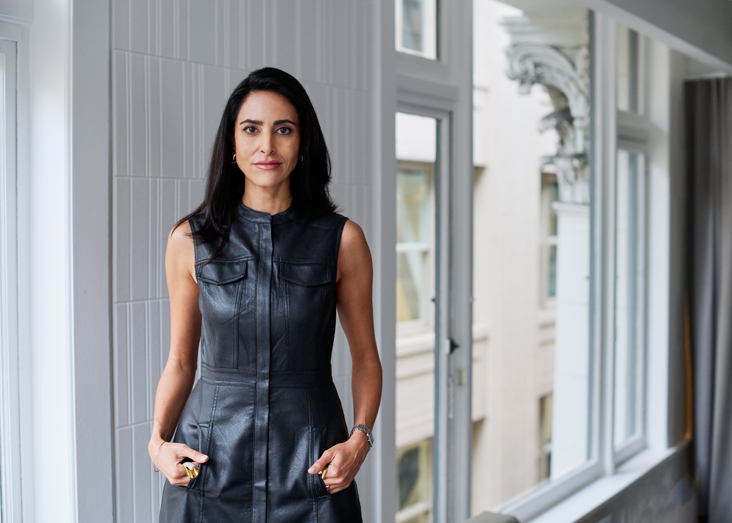 A woman with shoulder-length black hair wearing a sleeveless black leather dress standing in a bright indoor corridor with windows and decorative molding.