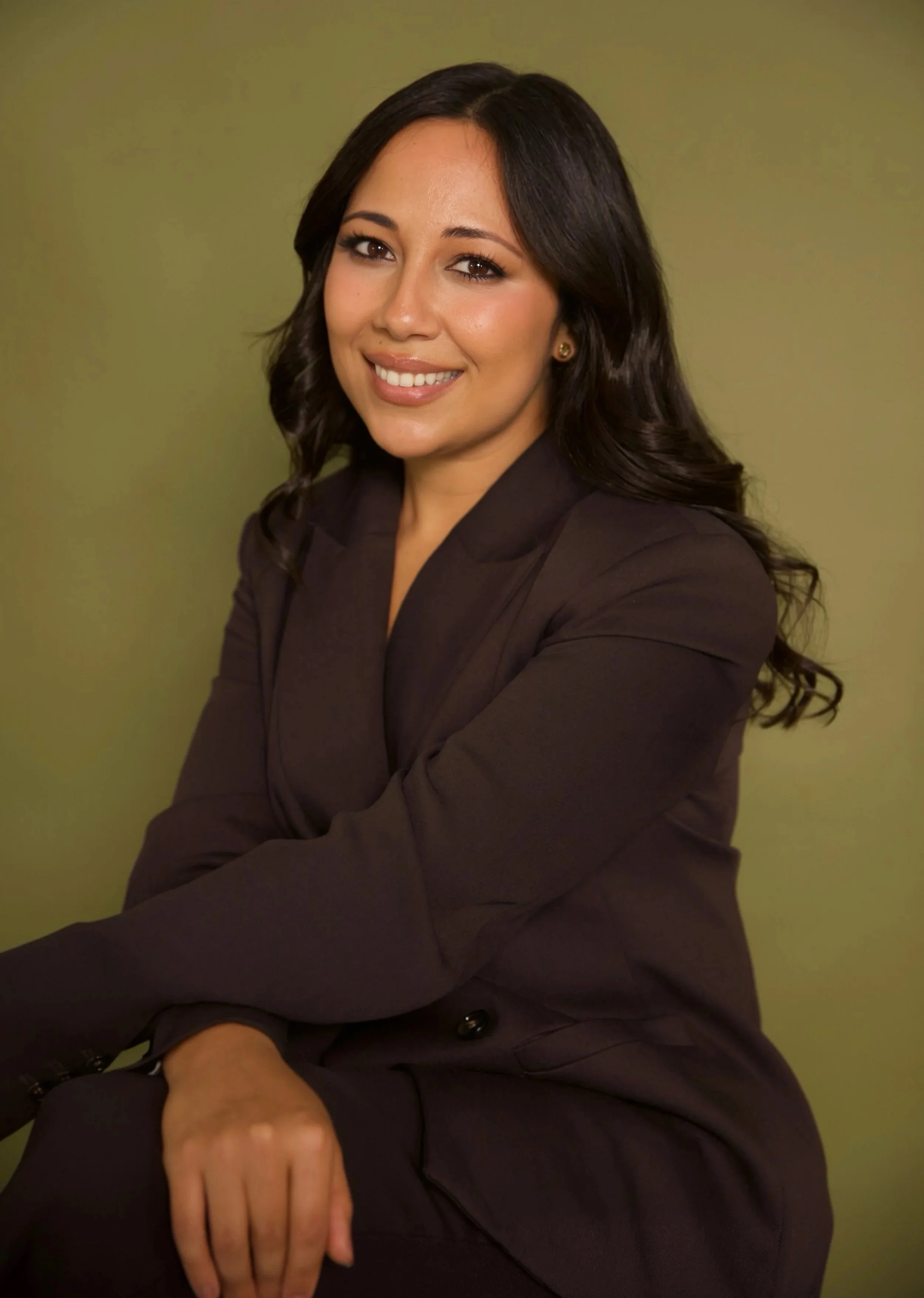 A young, female doctor with long dark wavy hair, smiling, and dressed in a brown blazer, sitting against a plain light green background.