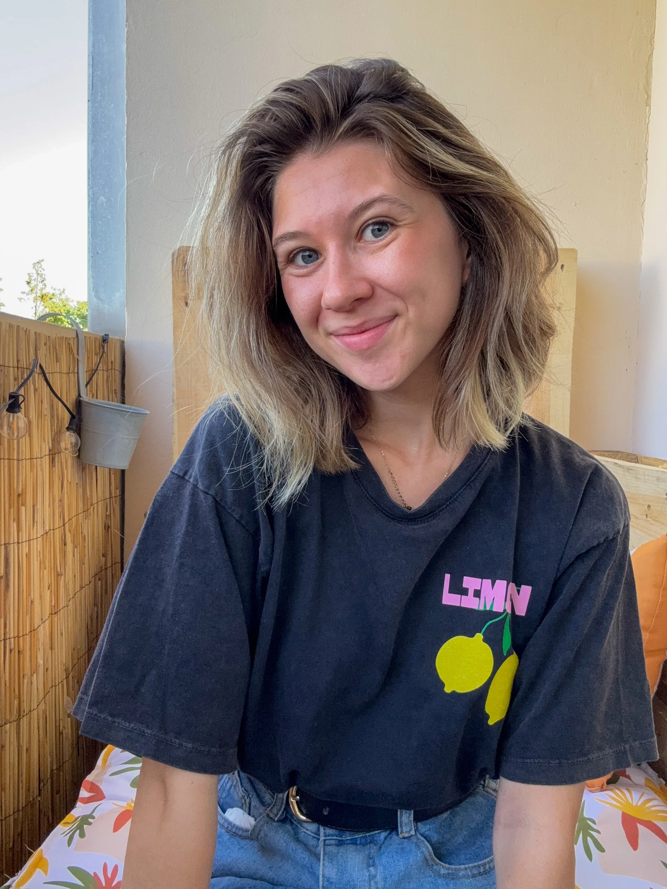 Jeune femme avec des cheveux bruns et blonds, portant un t-shirt noir avec un graphique de citrons, assise sur un lit avec une couverture colorée, dans une pièce ensoleillée avec un mur beige et un panneau en bambou. Explique son projet.