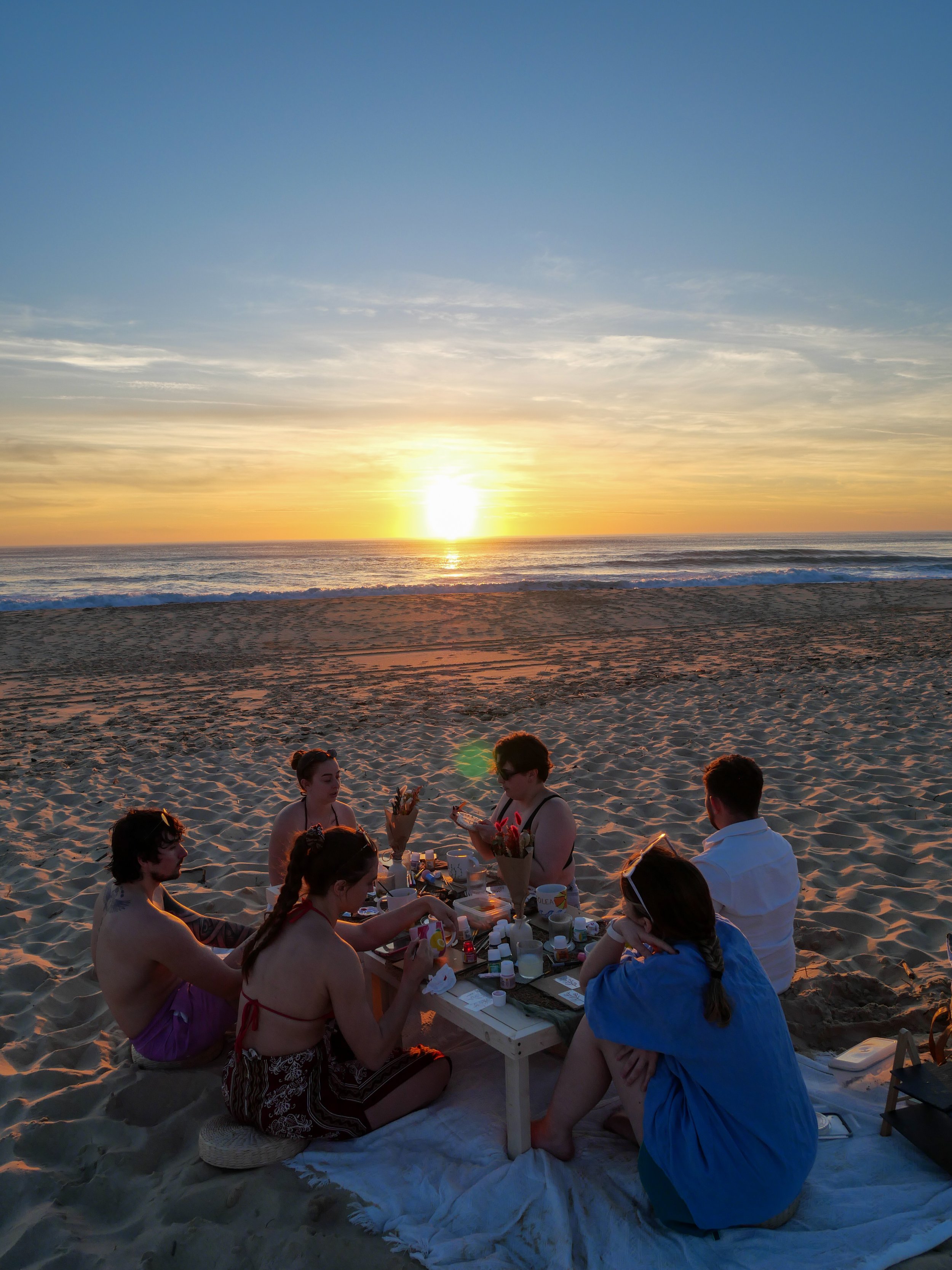 Groupe de personnes assises autour d'une table en bois sur la plage lors d'un coucher de soleil, partageant un repas ou des activités créatives. A partager, seul, entre amis, en couple en famille, pour un EVJF, un anniversaire...