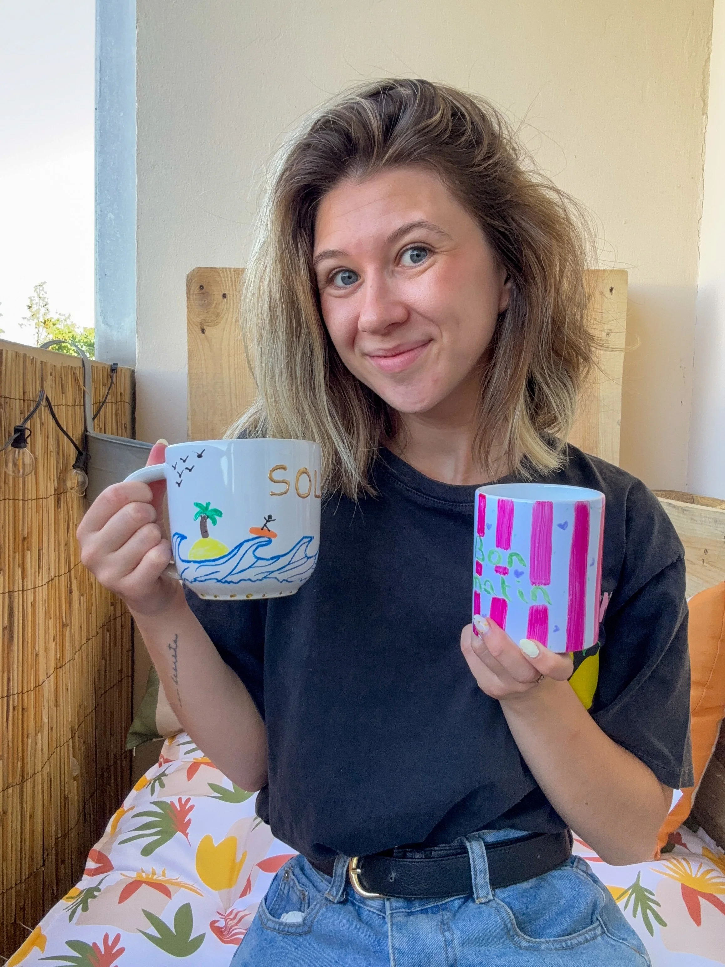 Une jeune femme souriante, tenant deux mugs décorés, assise sur une terrasse avec un panneau en bambou et un ciel clair en arrière-plan.