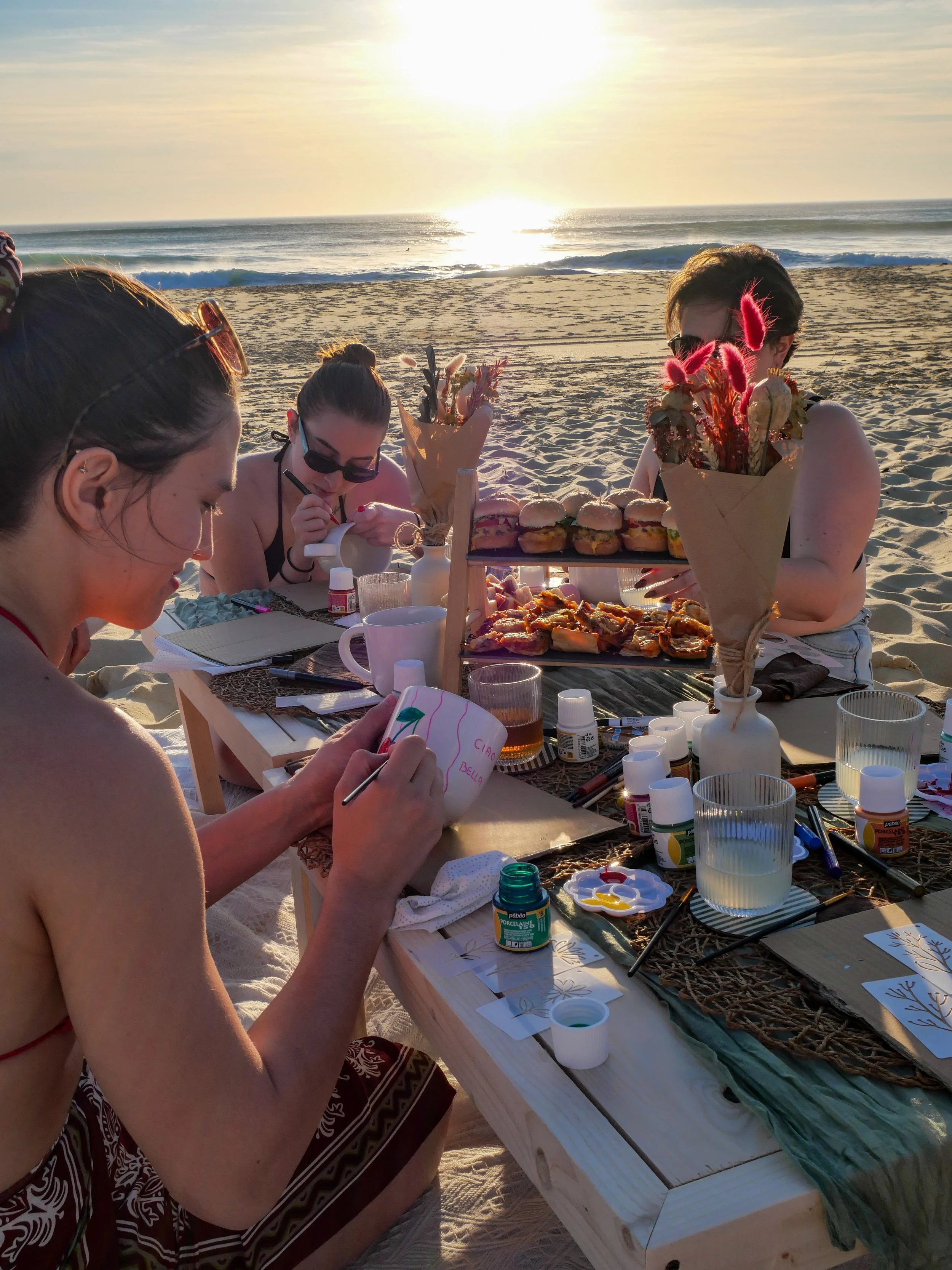 Groupe de personnes assises à une table sur la plage en train de peindre et de dessiner, avec des collations et des fleurs dans des bouquets en papier, lors du coucher du soleil.