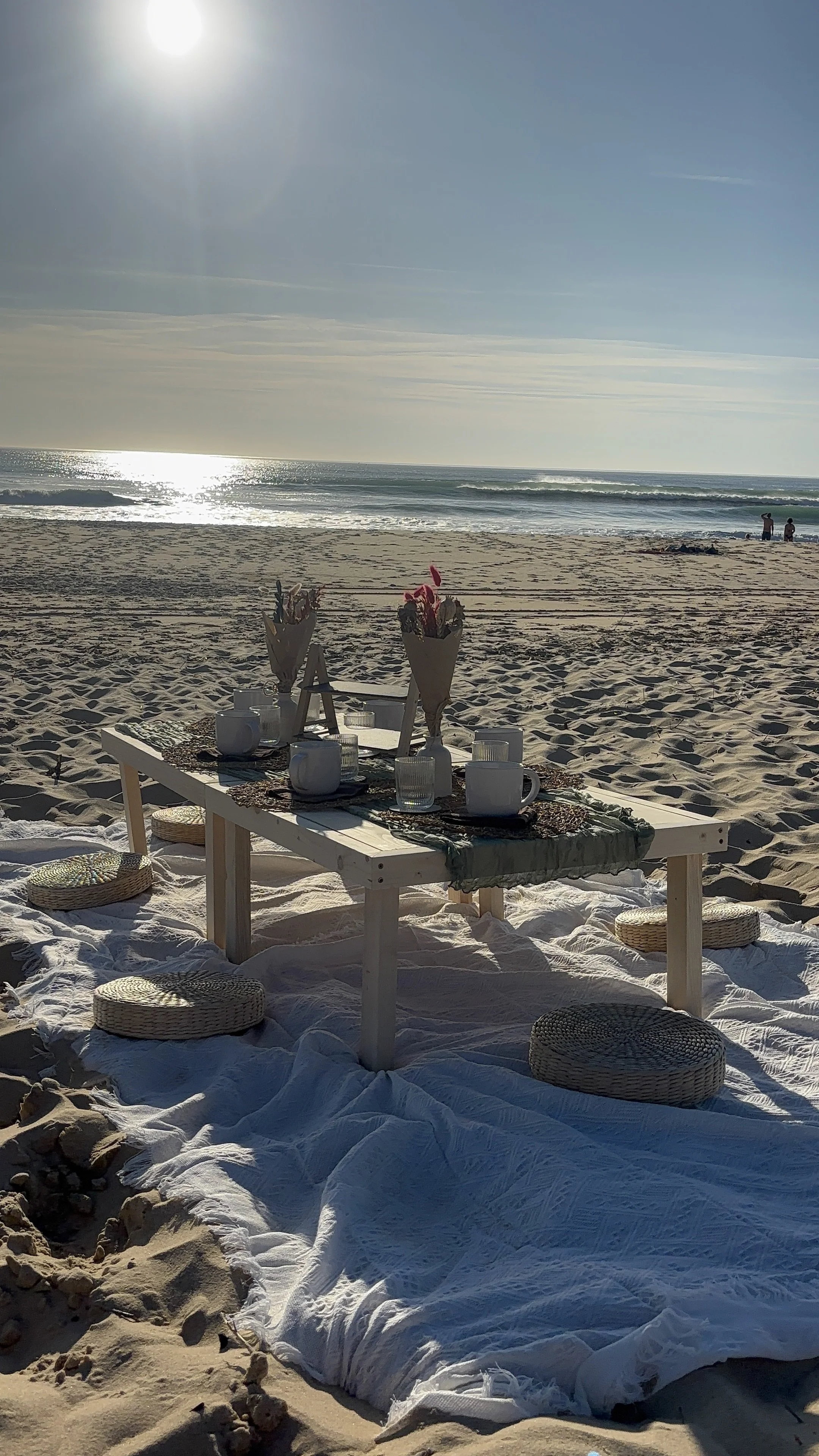 Table décorée avec des fleurs et des vaisselle, posée sur la plage face à la mer, avec des coussins en rotin autour sur le sable, sous un ciel ensoleillé.