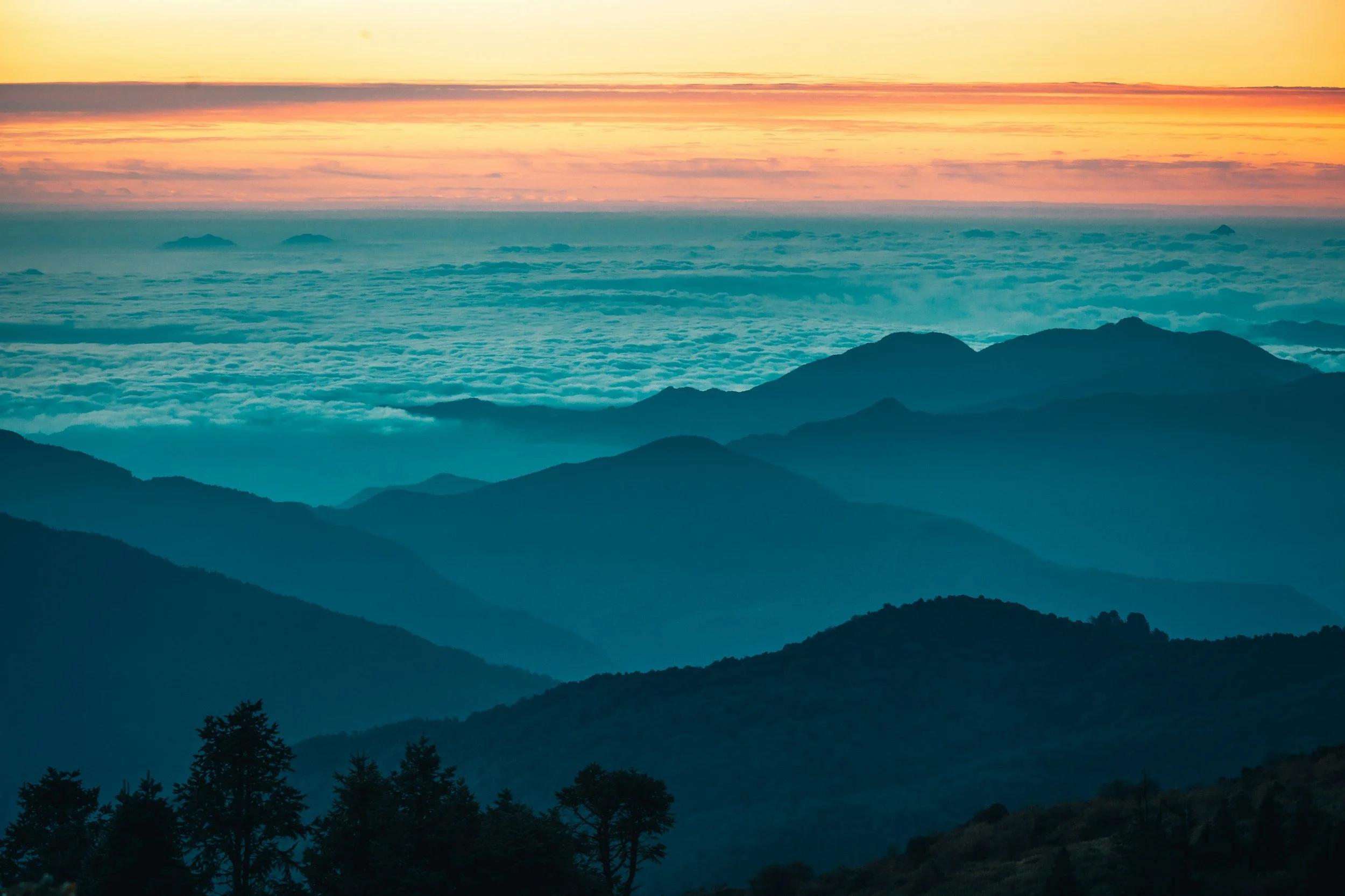 Sunrise over mountain range with layers of clouds and silhouetted trees in the foreground.