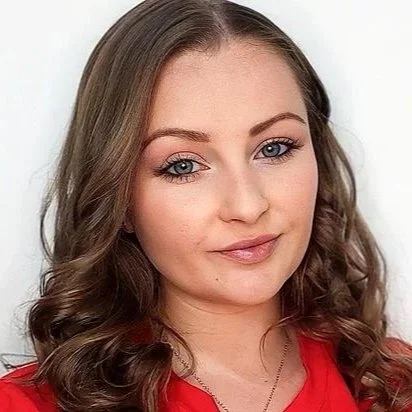 Close-up of a young woman with long, wavy brown hair and blue eyes, wearing a red top and smiling softly.
