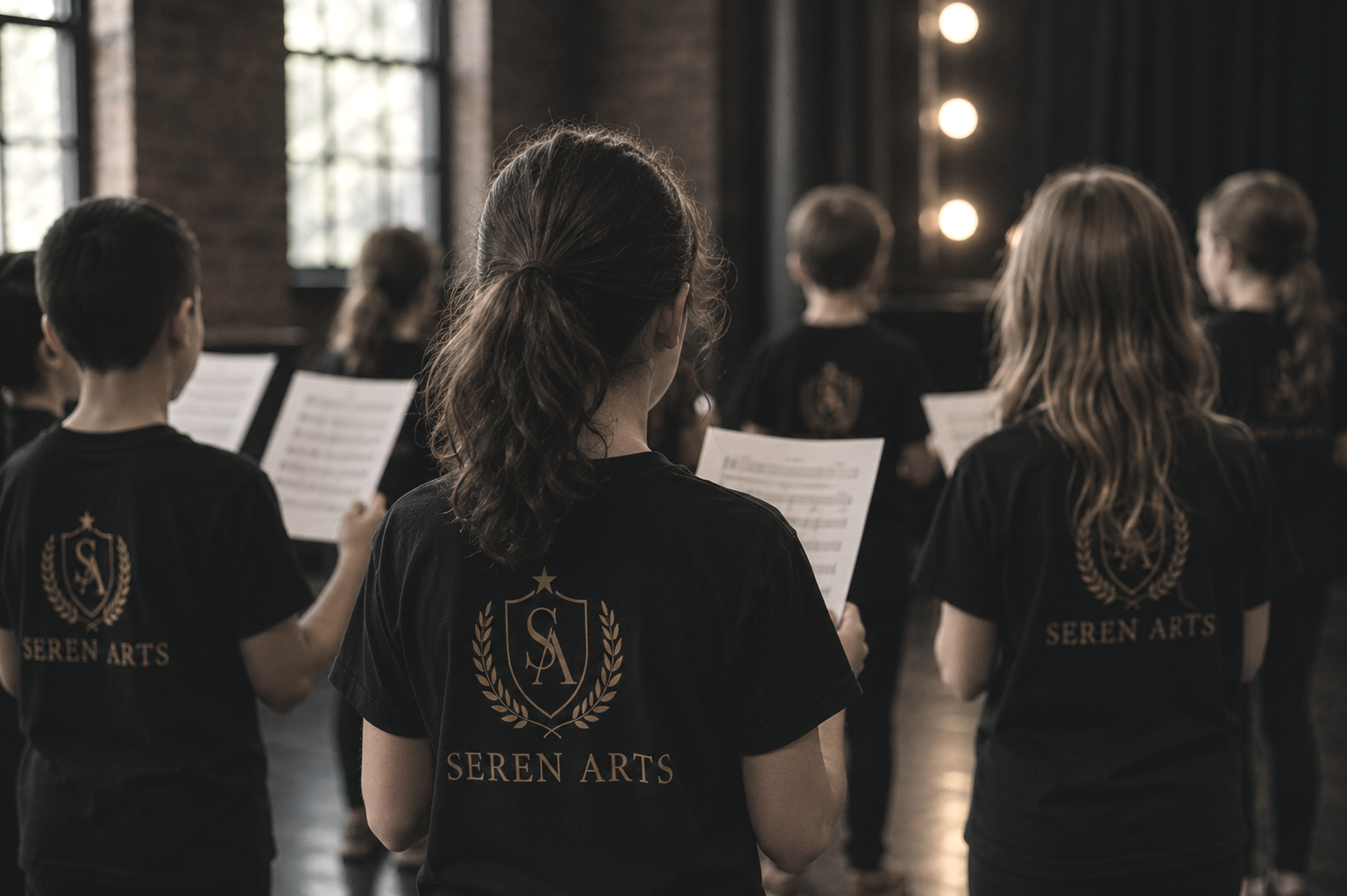 Group of children wearing black shirts with 'Seren Arts' logo, practicing or performing music with sheet music in a rehearsal space with large windows and brick walls.